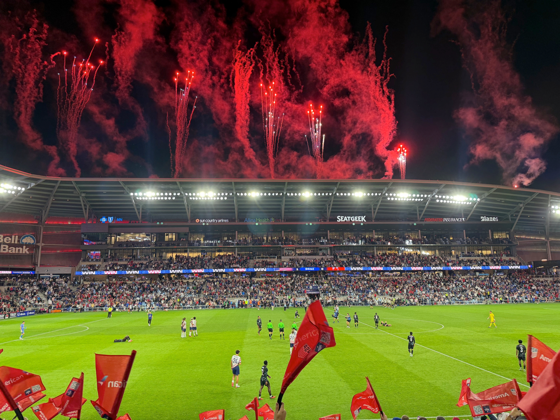 Red fireworks burst above the stadium roof as players walk the field and fans below wave red flags.