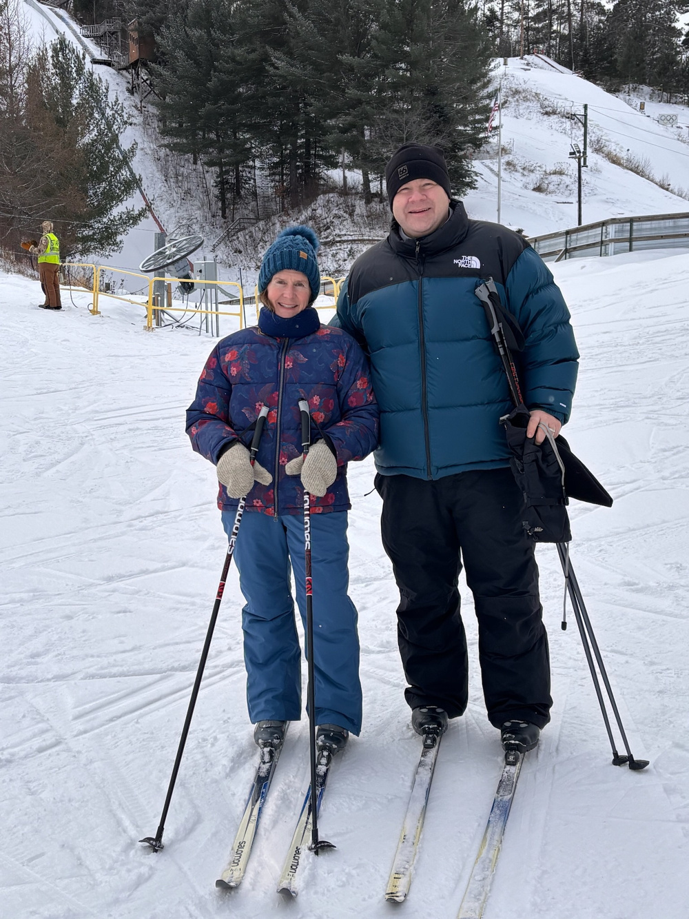 Two people are smiling and posing with skis on a snowy slope, surrounded by trees and ski equipment.