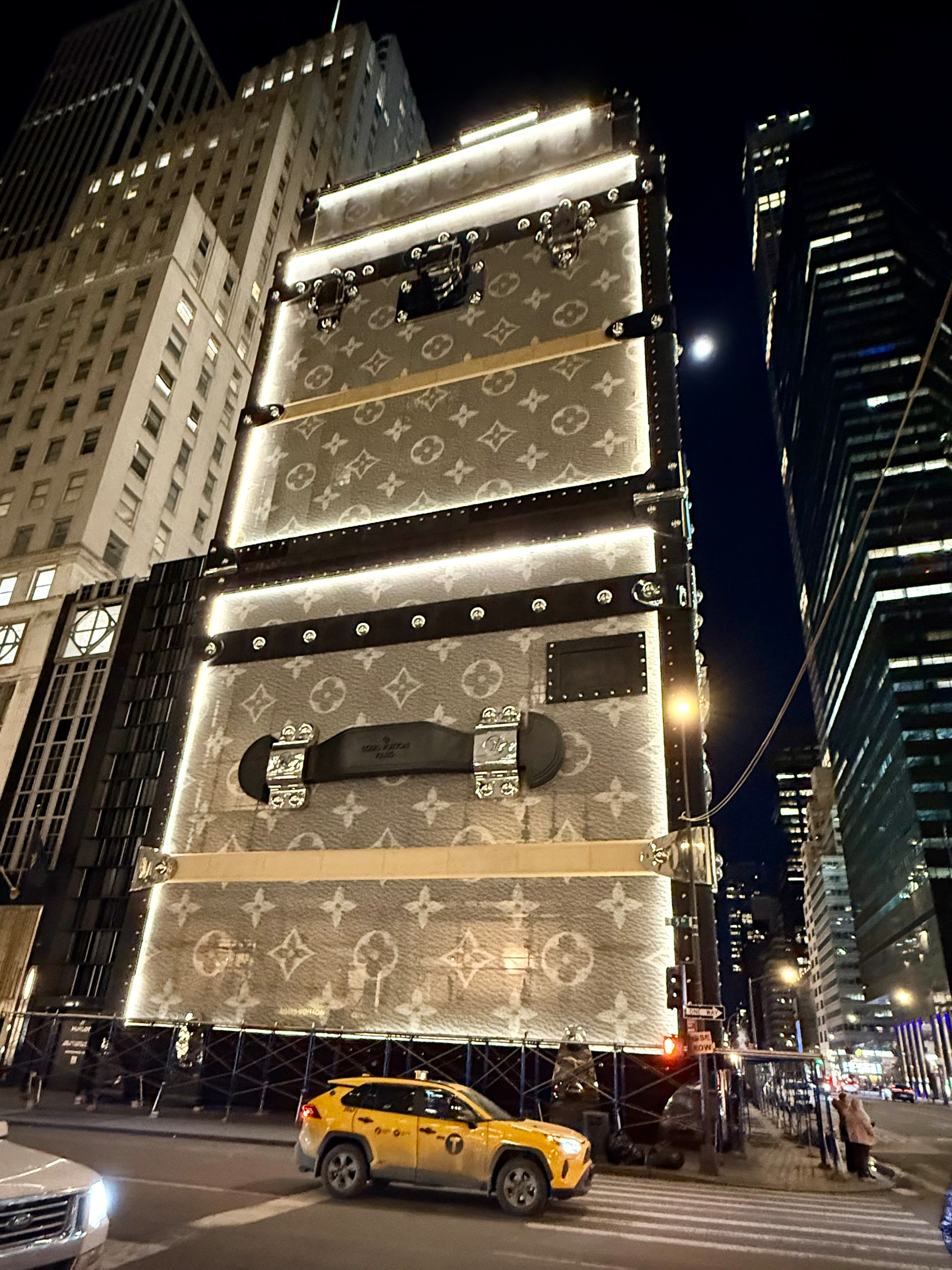 Auto-generated description: A giant, illuminated Louis Vuitton trunk, adorned with monogram patterns, dominates a city street at night.