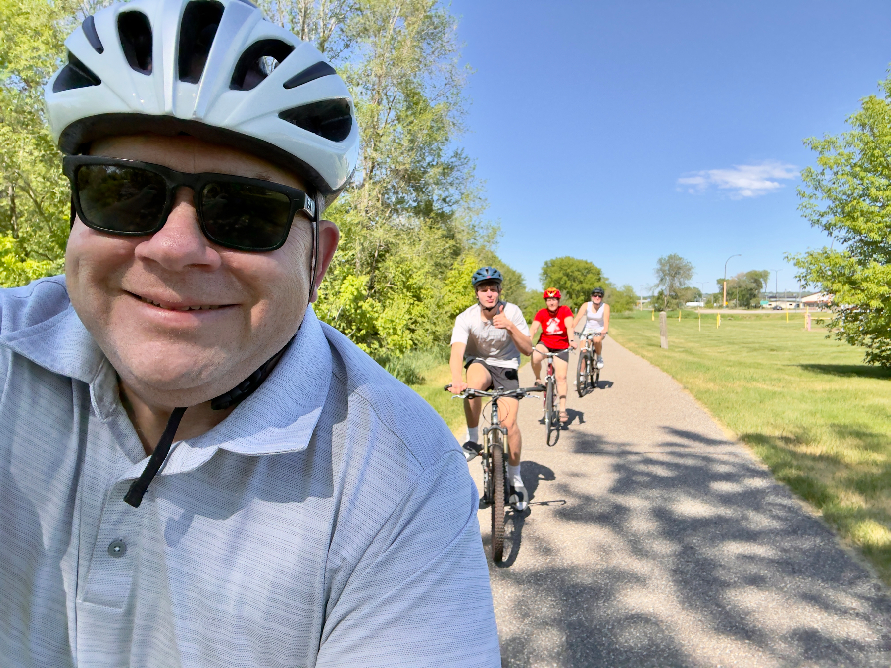 Man in white helmet and sunglasses takes a selfie on a paved trail while three cyclists ride behind him on a sunny day.