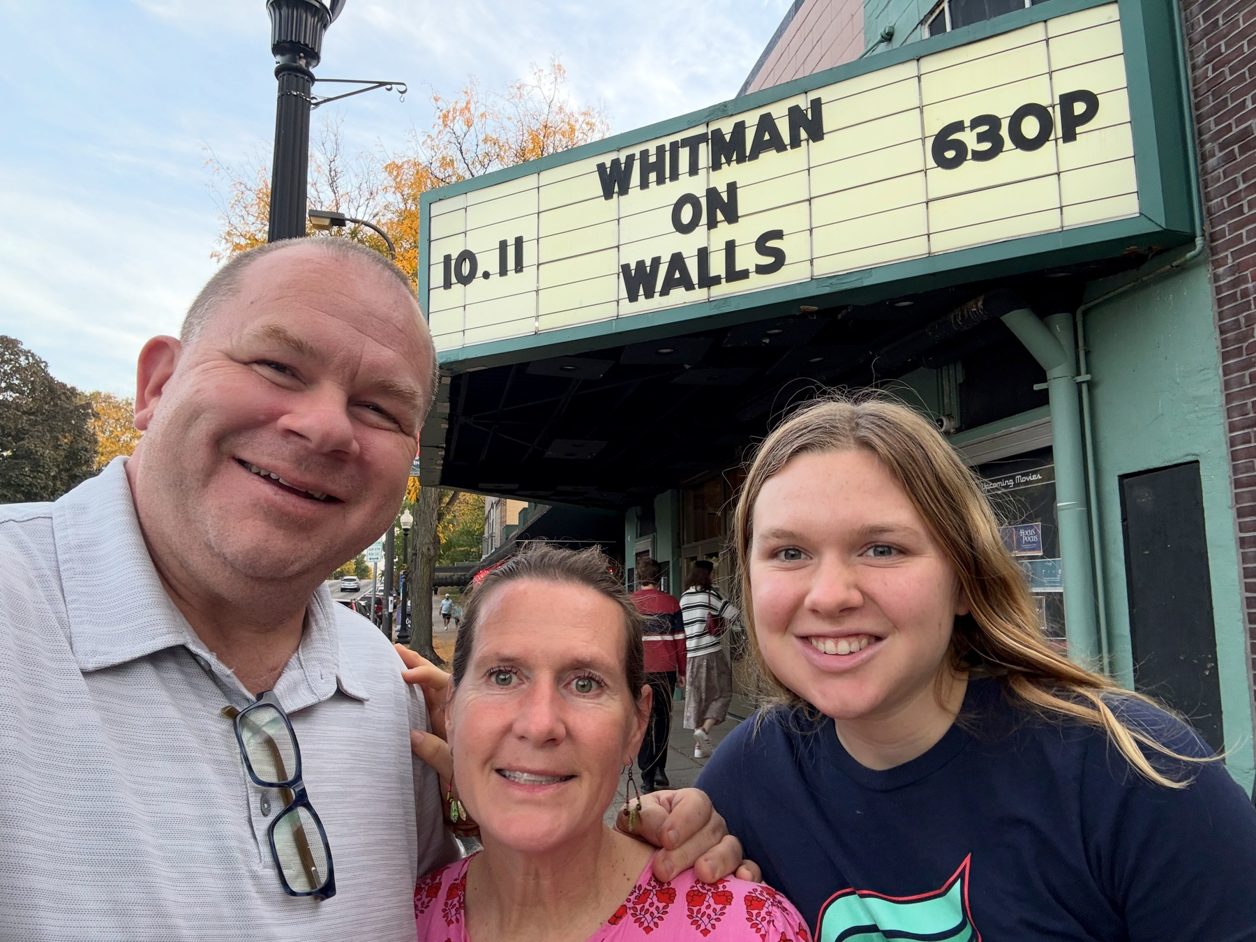 Selfie of a man and two women under a theater marquee reading 10.11 Whitman On Walls 630P, autumn trees behind them.