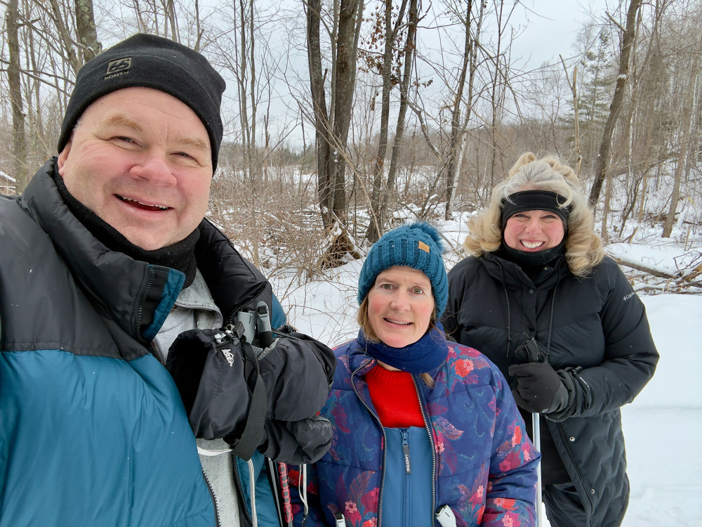 Three people dressed in winter clothing are smiling while standing in a snowy, wooded area.