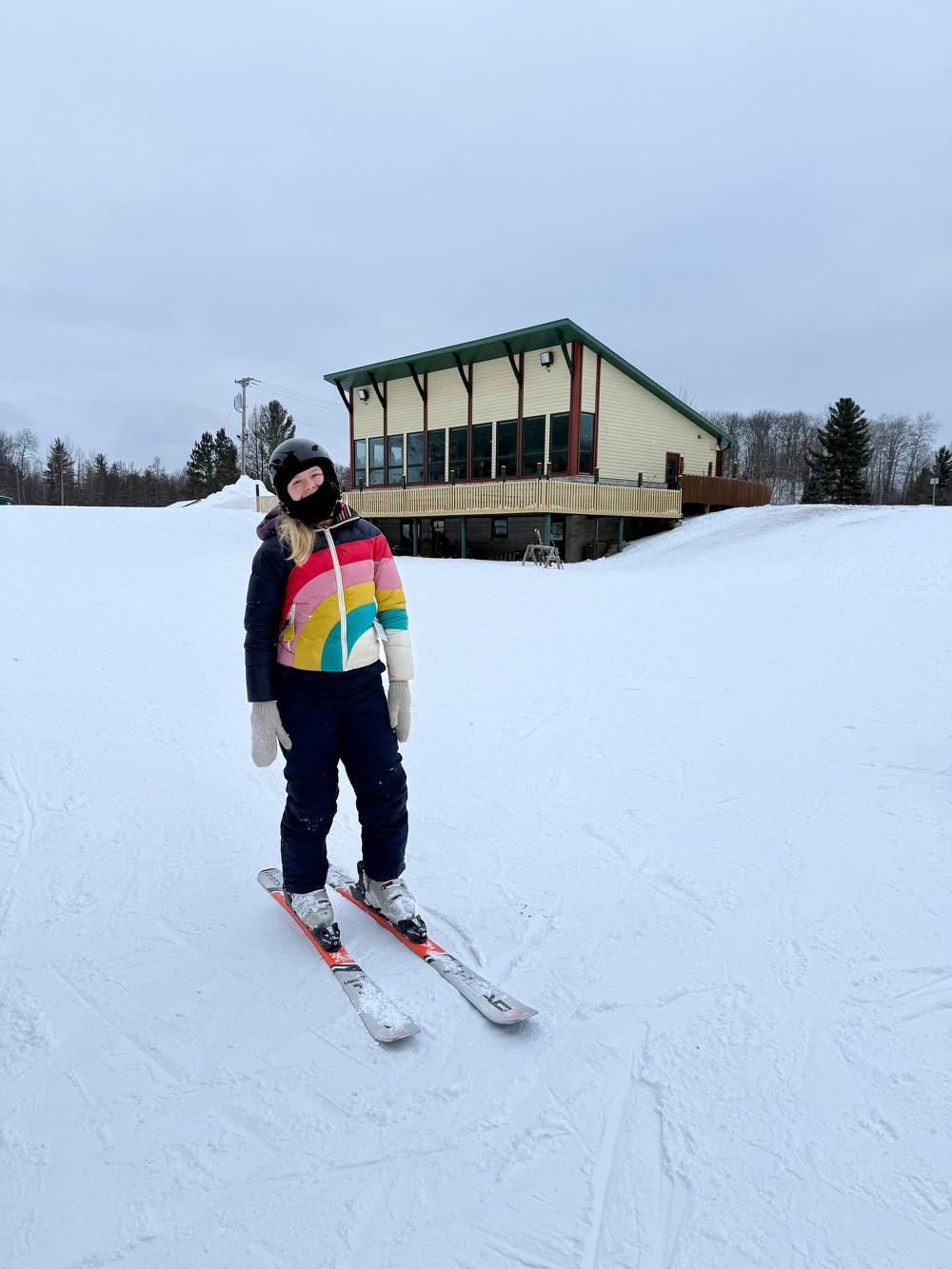 A person wearing a colorful jacket is standing on skis in a snowy area with a building in the background.