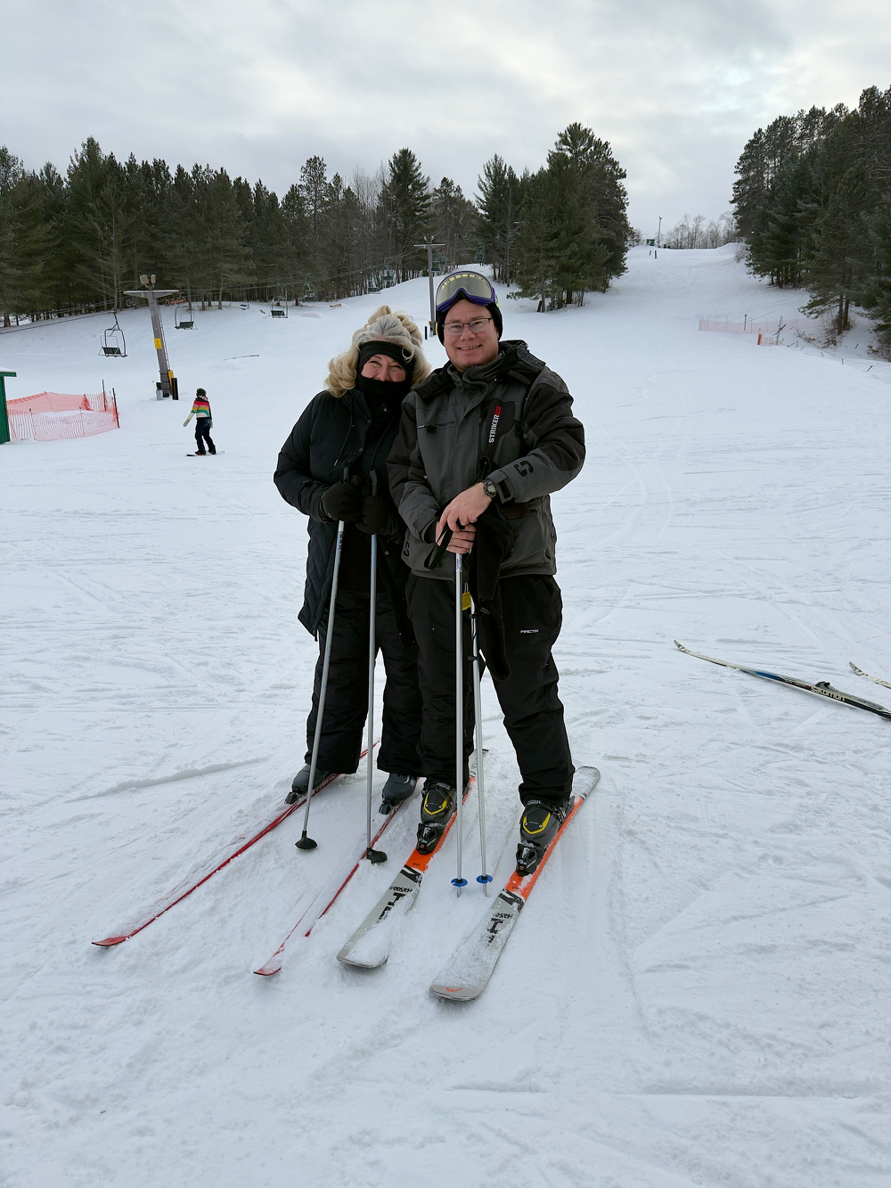 Two people in winter clothing are skiing on a snowy slope, surrounded by trees and a ski lift.