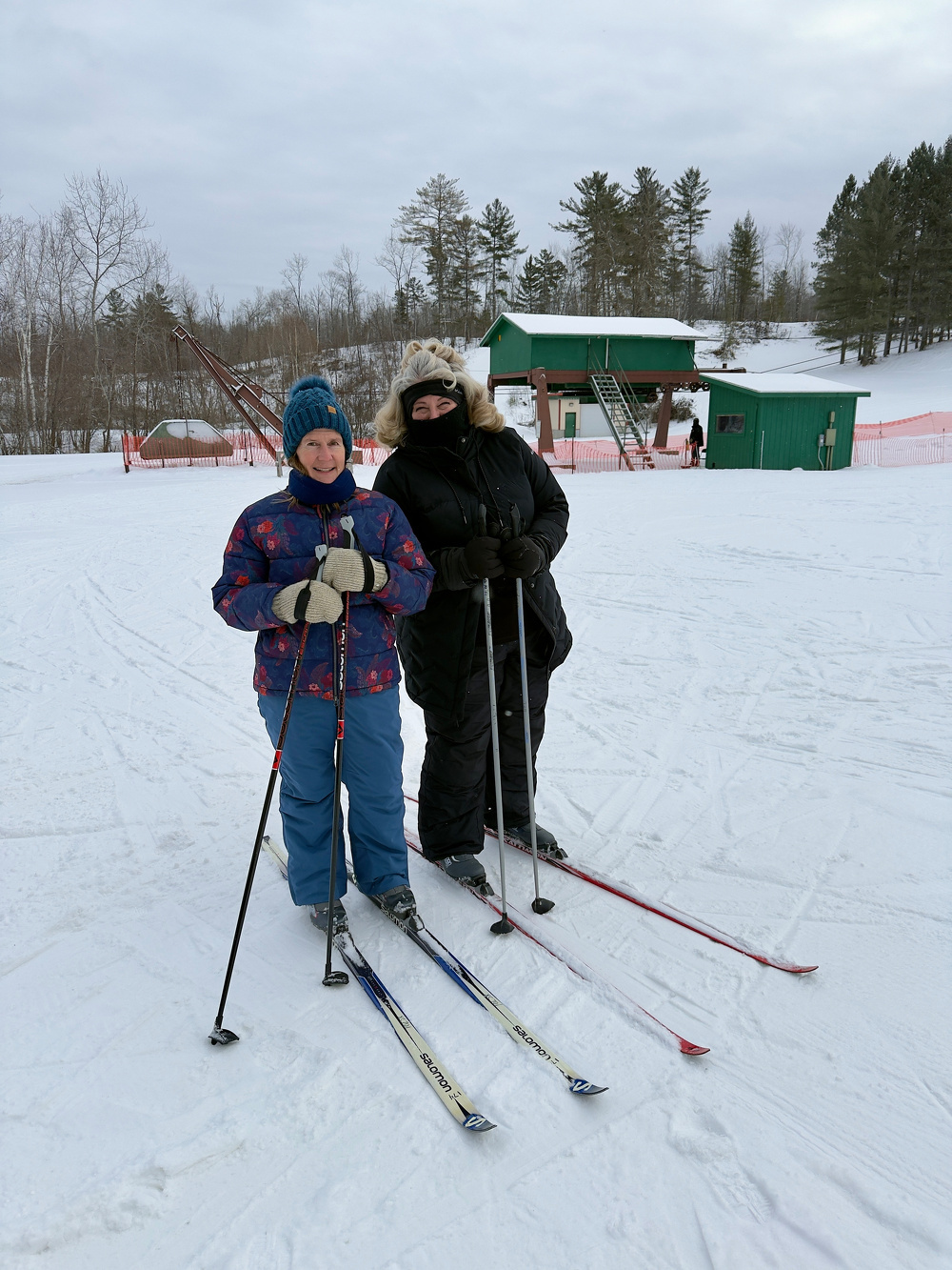 Two people dressed in winter clothing are standing on a snowy field with ski equipment, with a green building and trees in the background.