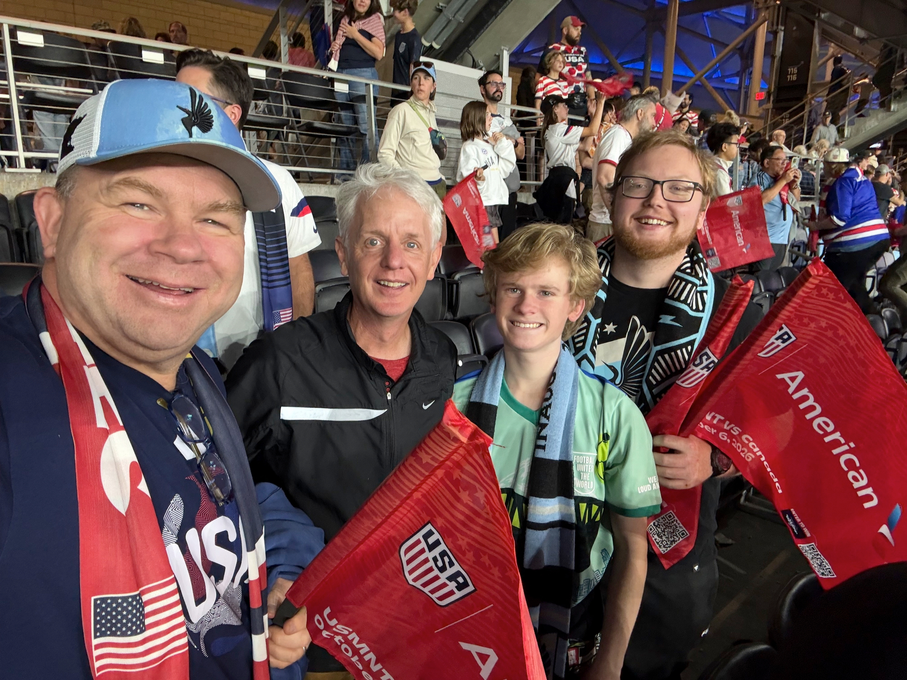 Four people in soccer scarves, two men, a teen, and a young man, pose for a selfie in the stands holding red USA flags.
