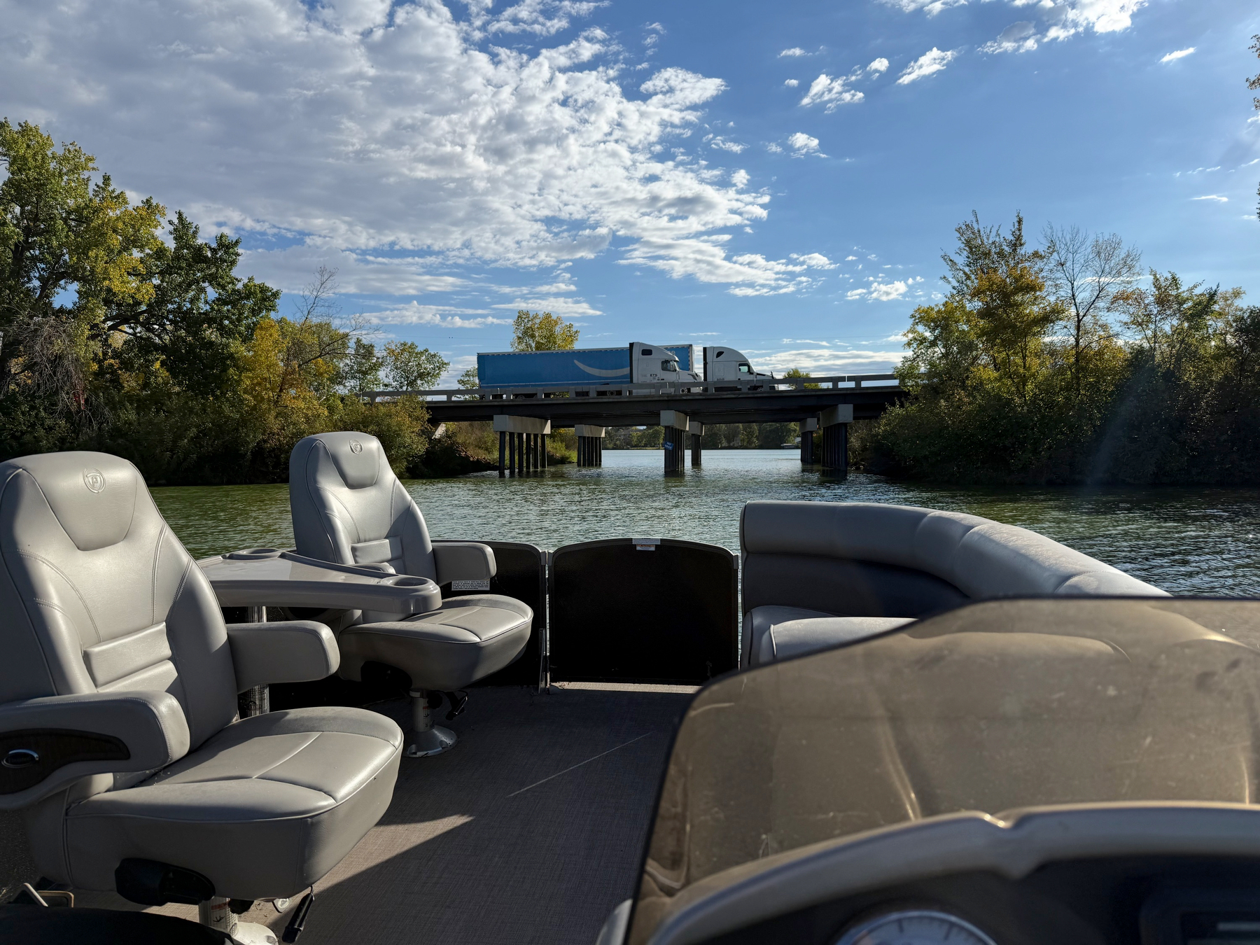 A pontoon's gray seats face a low bridge over a tree-lined channel, where two semi trucks are crossing.
