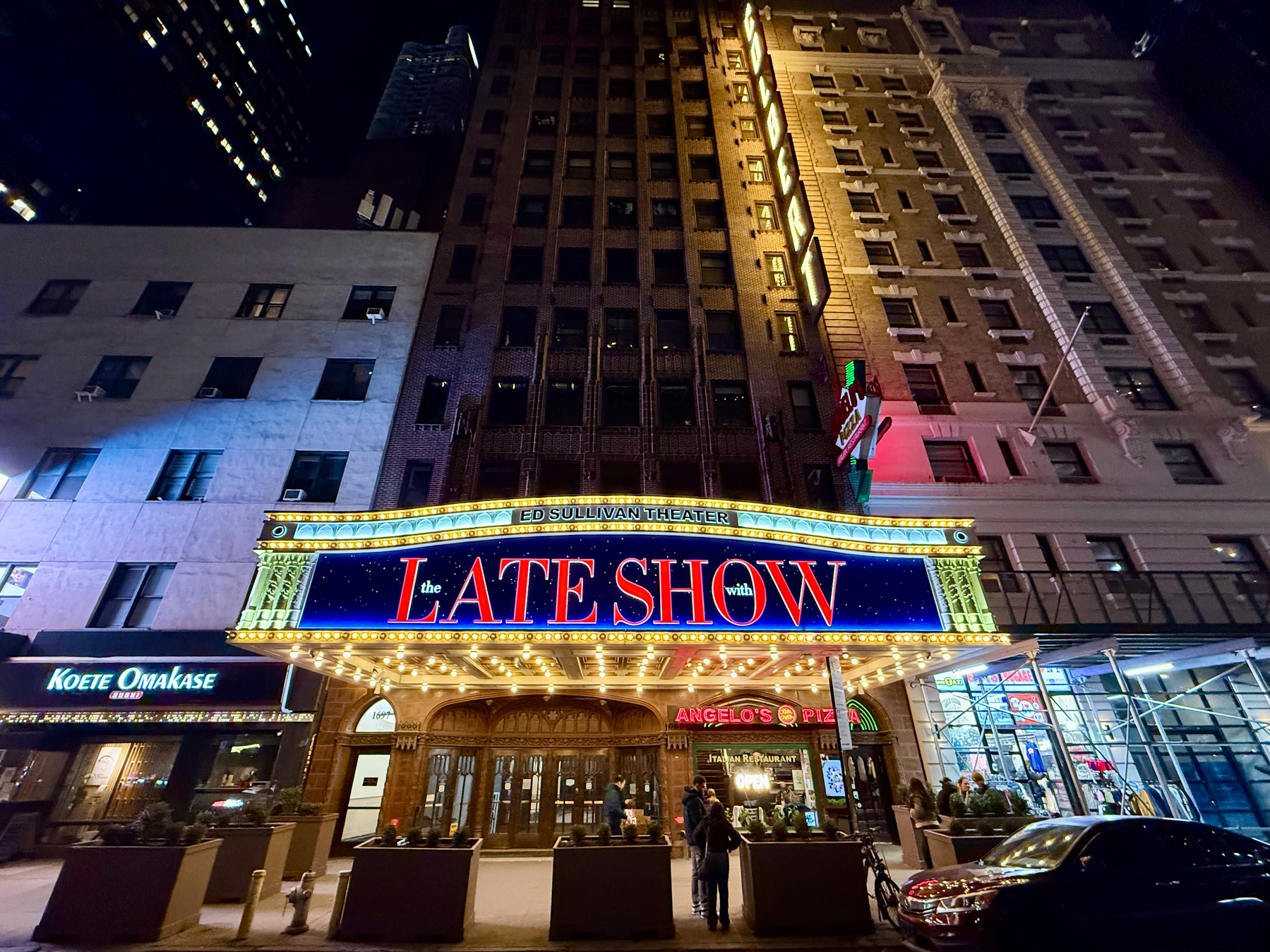 Auto-generated description: A brightly lit theater marquee for The Late Show is displayed on a city street at night.