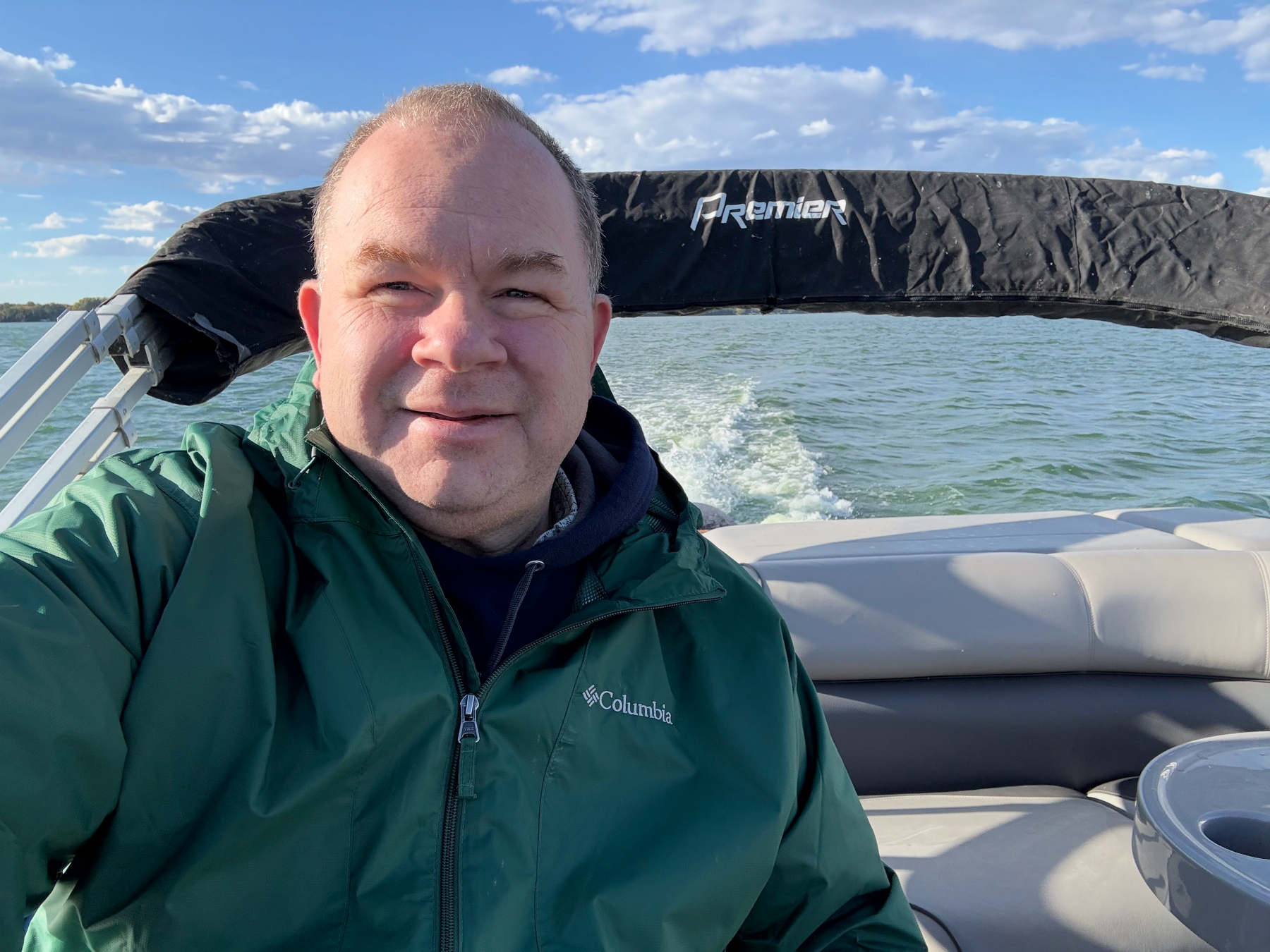 A man in a green rain jacket takes a selfie at the back of a pontoon boat, its wake trailing across open lake water.