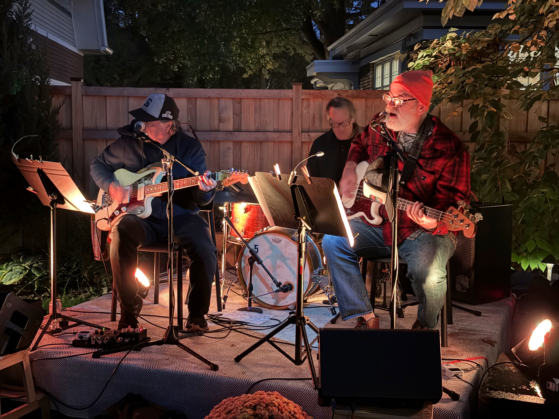 Guitarist in a cap, drummer, and singing bassist in red plaid play on a lit backyard stage at night before a wooden fence.