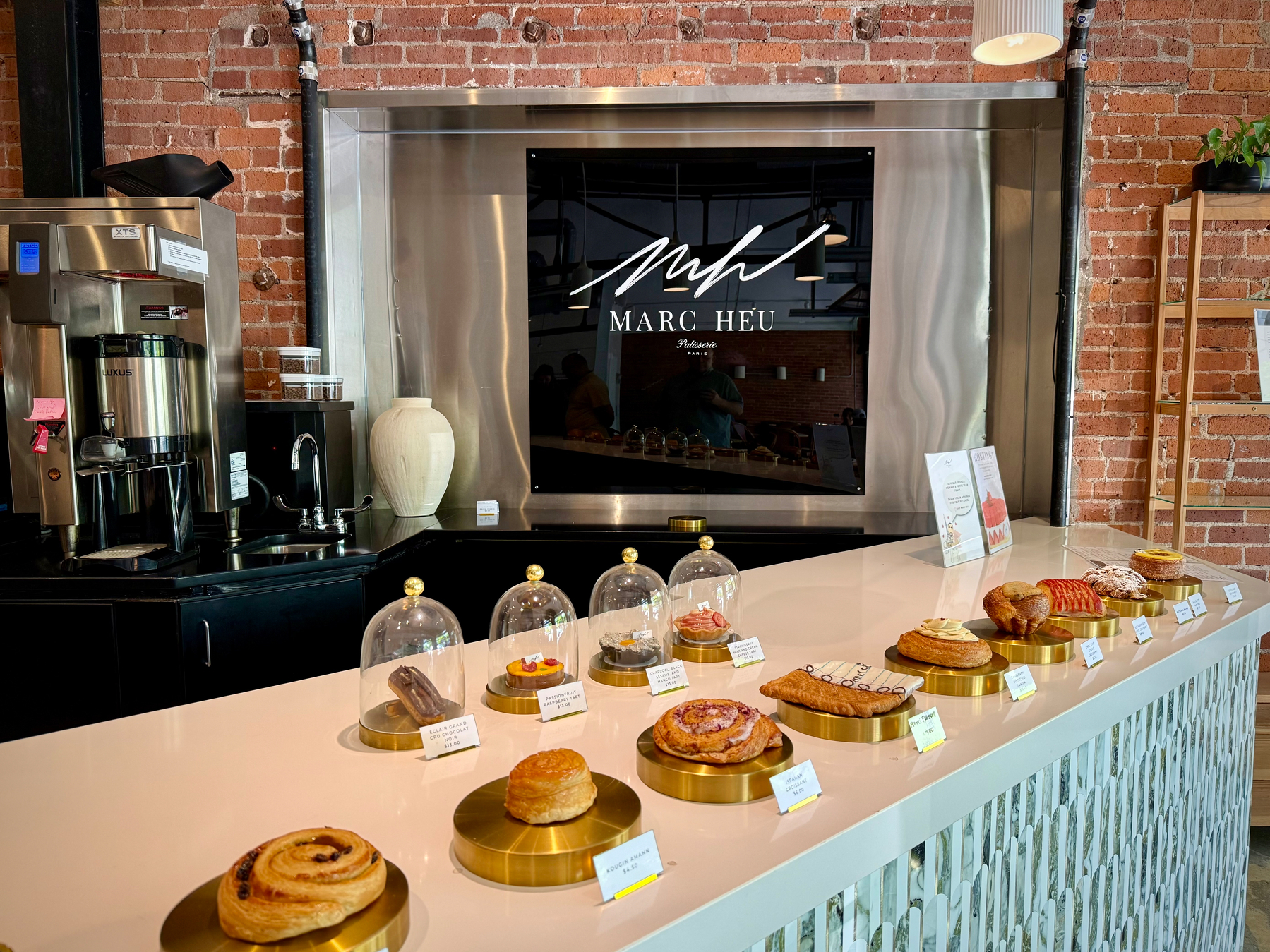 Display counter at Marc Heu Patisserie showing pastries and tarts on gold stands beneath glass domes, with brick walls and espresso equipment behind.