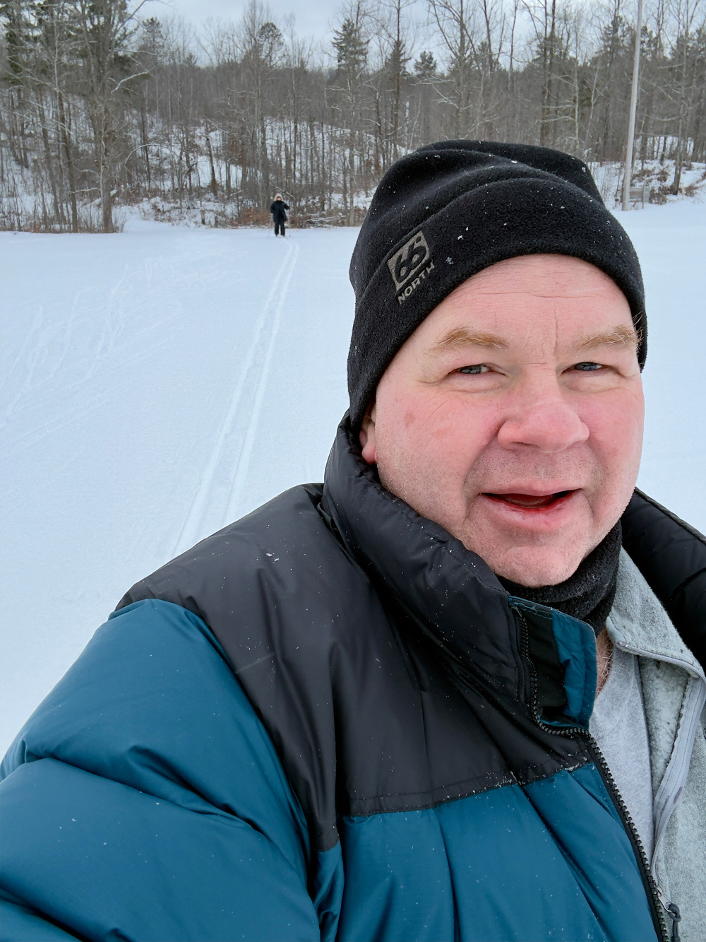 A person in a winter jacket and beanie takes a selfie in the snow with another person in the distance.