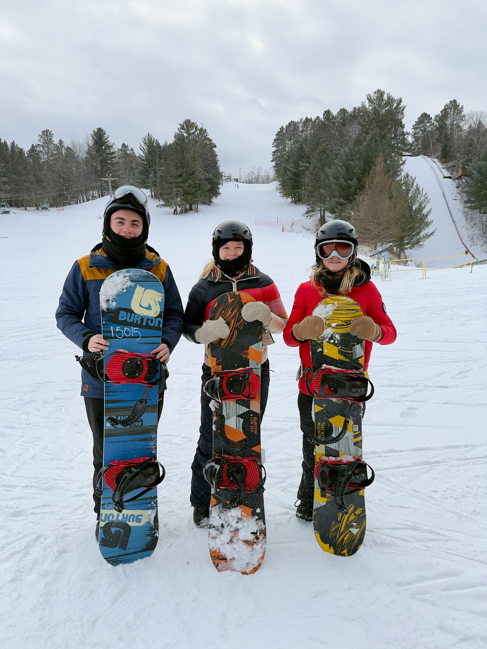 Three snowboarders wearing helmets pose with their snowboards on a snowy slope surrounded by trees.