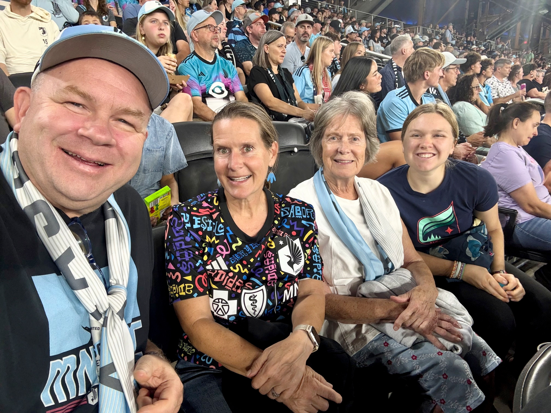 Selfie of a man in a cap and scarf with three women seated in a crowded stadium, fans in light blue MNUFC jerseys behind.