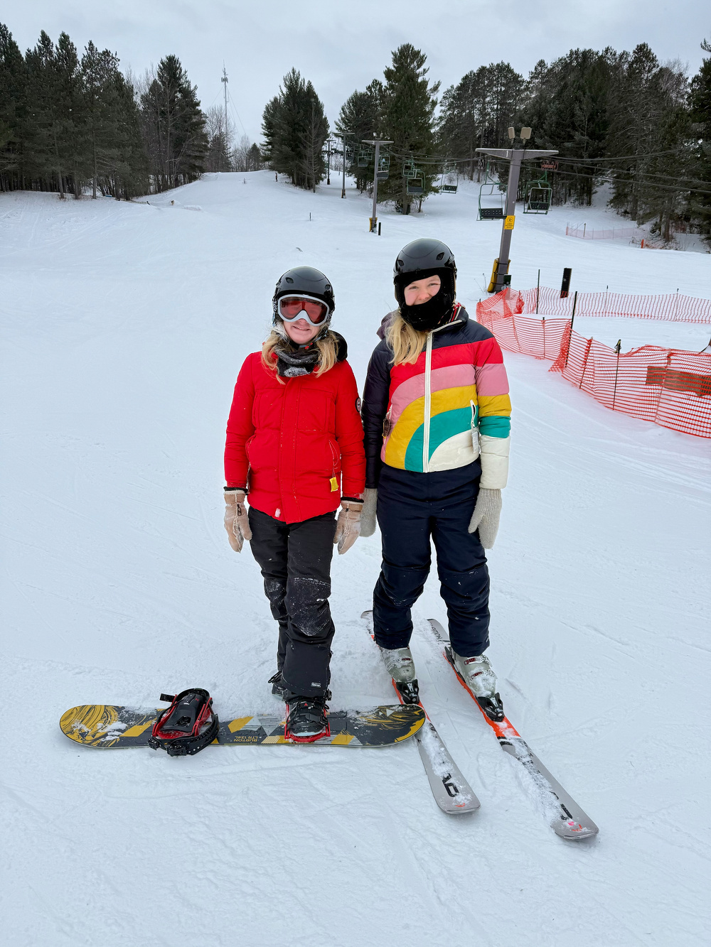Two people in winter gear stand on a snowy ski slope, with one wearing a red jacket and snowboard, and the other in a colorful jacket with skis.