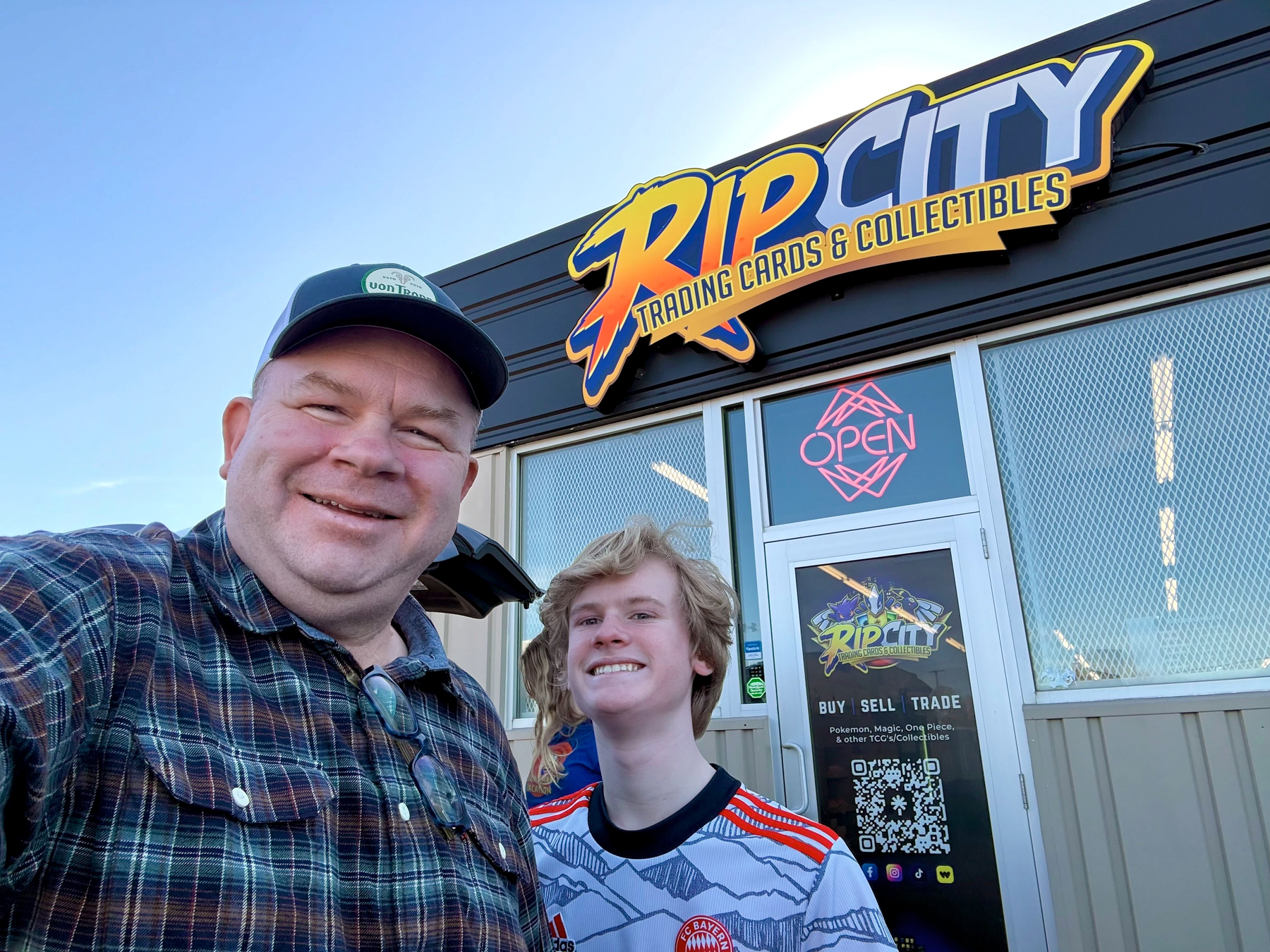 Selfie of a man in a plaid shirt and cap beside a teen in a Bayern jersey outside a trading card store with an open sign.