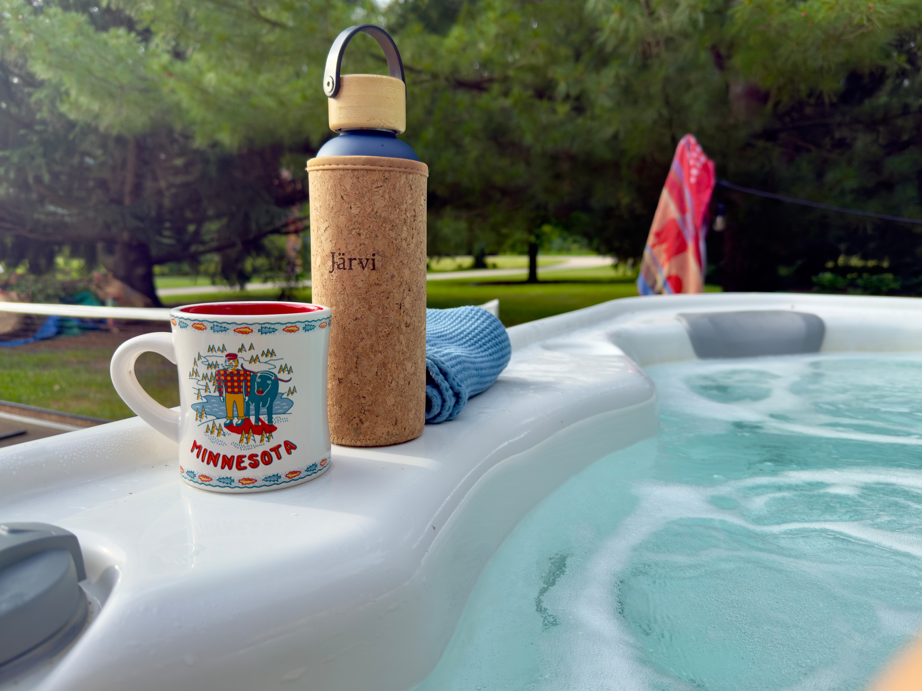 Minnesota souvenir mug and cork-sleeved Jarvi water bottle resting on the edge of a bubbling outdoor hot tub surrounded by trees.