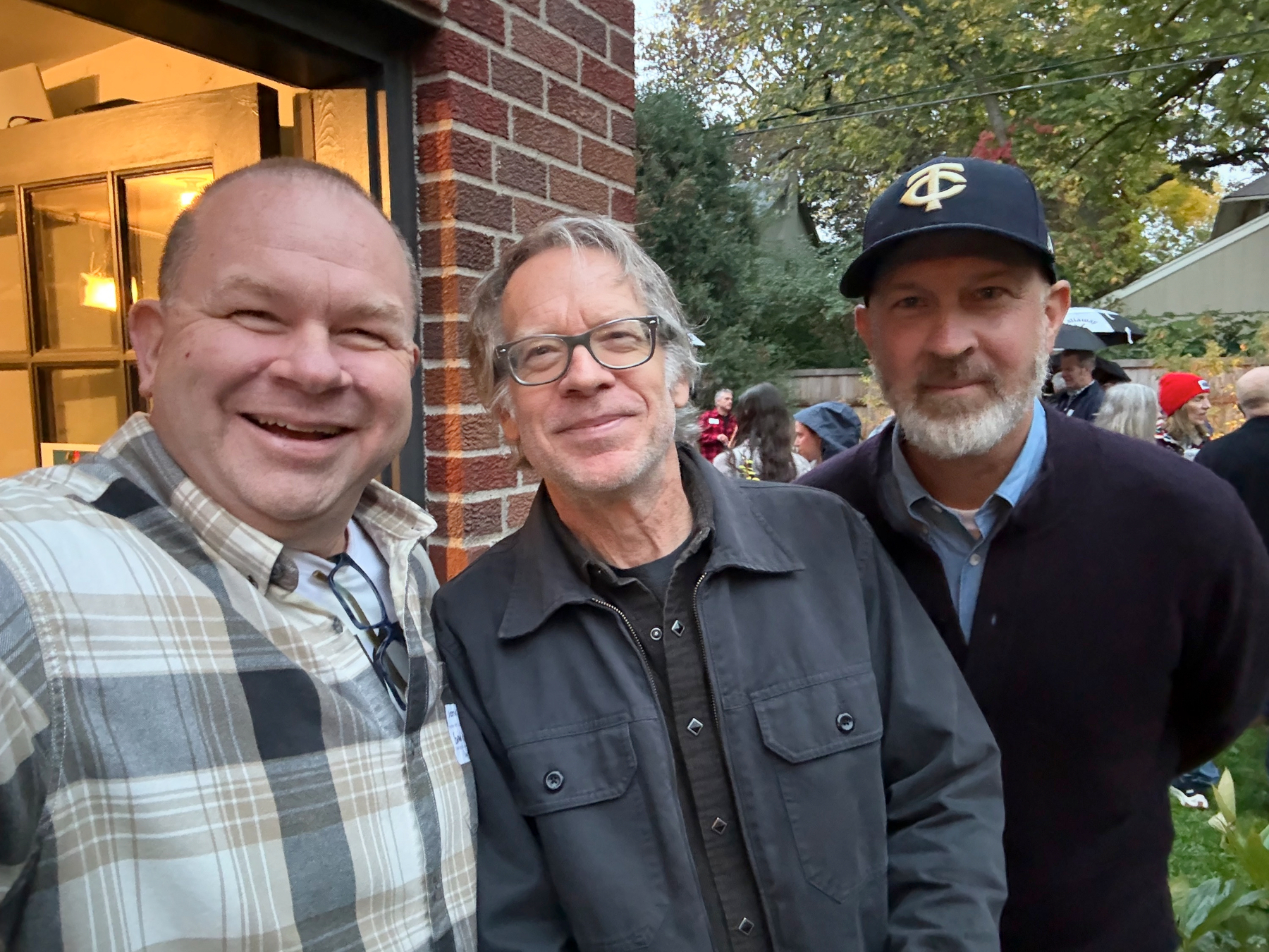Three men pose for a selfie beside a brick house at a backyard gathering; one wears glasses, another a Twins cap.