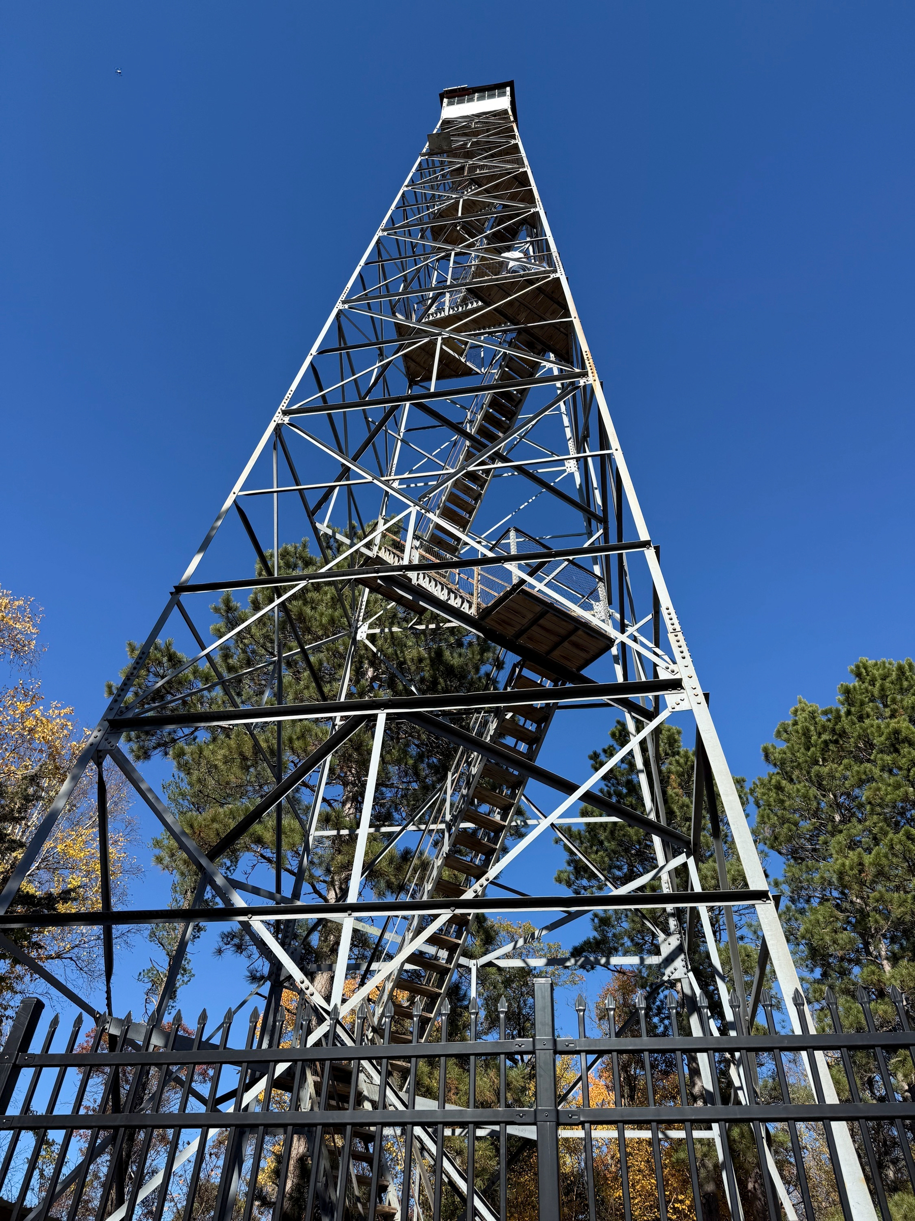 Steel lattice tower with switchback wooden stairs rising to a small cab, seen from below behind a black iron fence and pines.