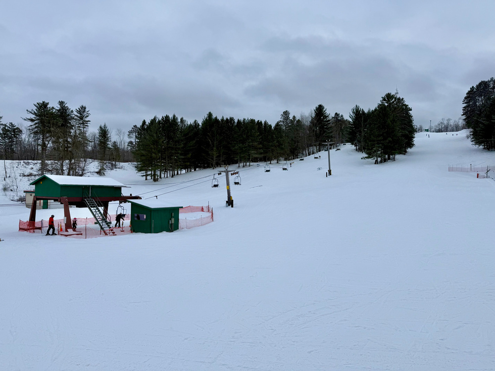 A snowy ski slope features a green ski lift station and is surrounded by trees under a cloudy sky.