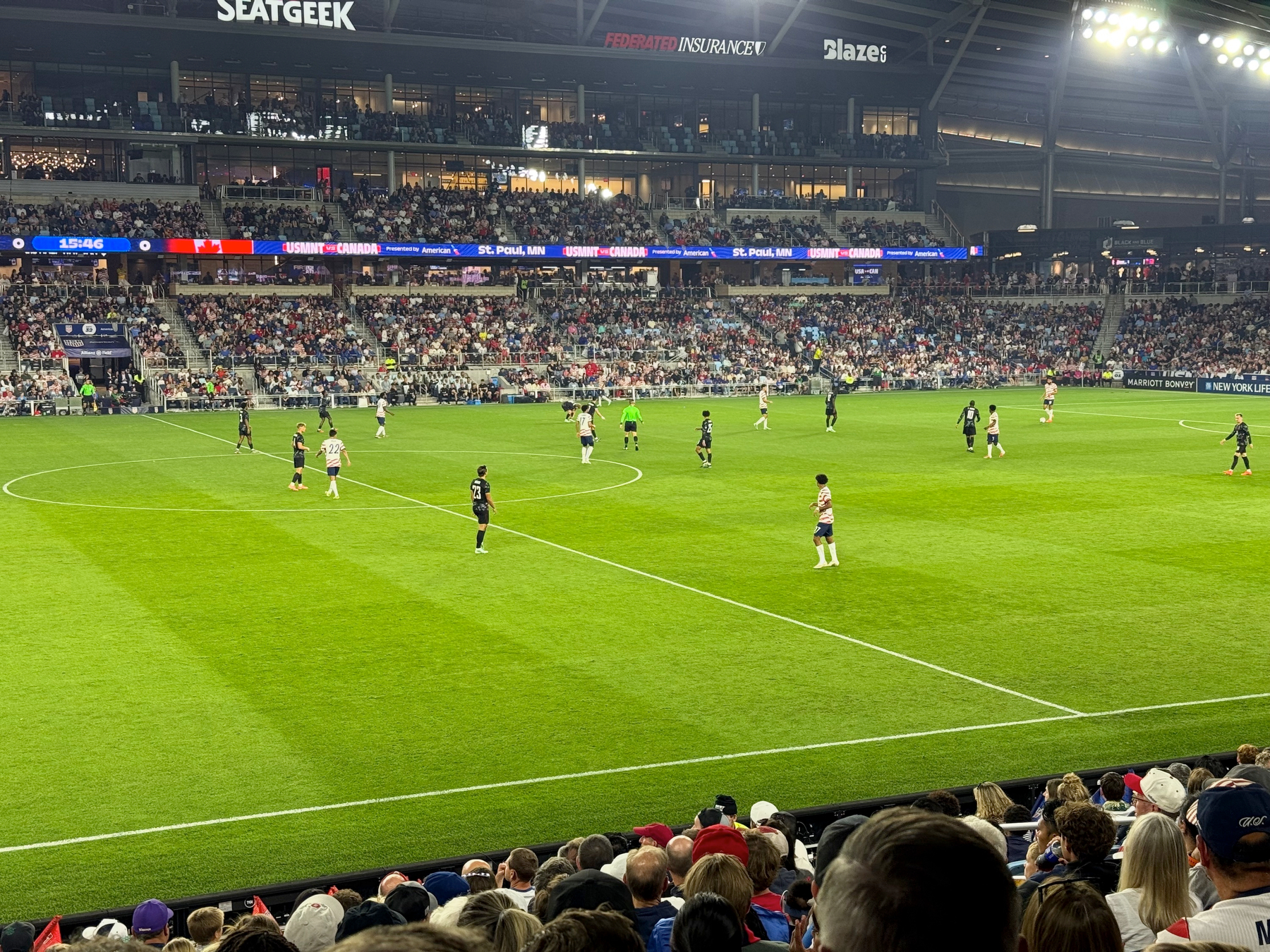 Players in white and black kits spread across a lit soccer pitch near midfield, with full stands and a ribbon board behind.