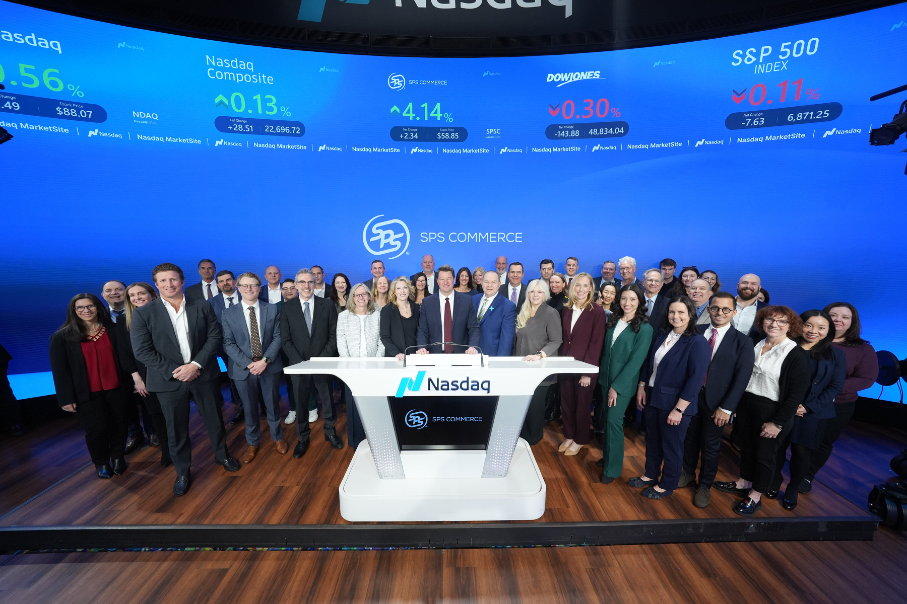 A large group of people poses around the Nasdaq podium on a stage with stock market displays in the background.