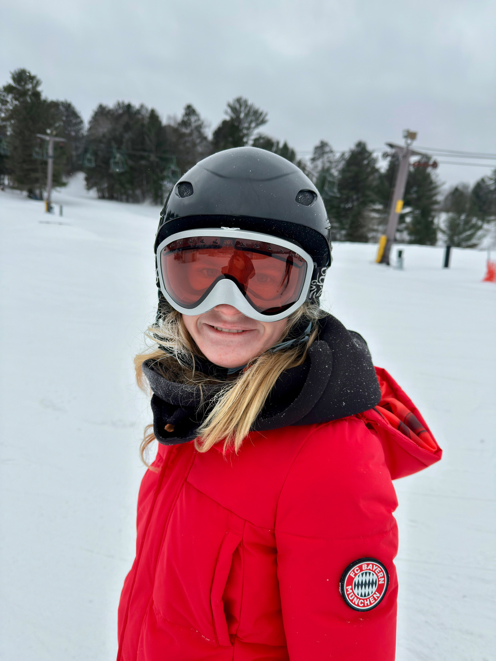 A person wearing a red winter jacket, ski helmet, and goggles is smiling on a snowy landscape with trees and a ski lift in the background.