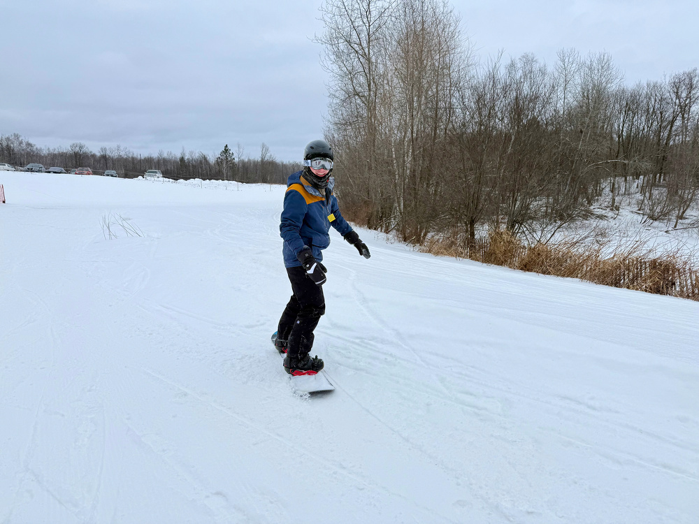 A person is snowboarding down a snowy slope with trees in the background.