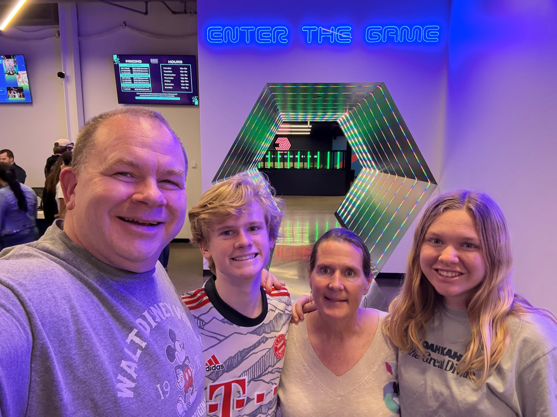 Selfie of four people under a blue neon "Enter the Game" sign, with a lit hexagonal tunnel entrance and pricing screen behind.