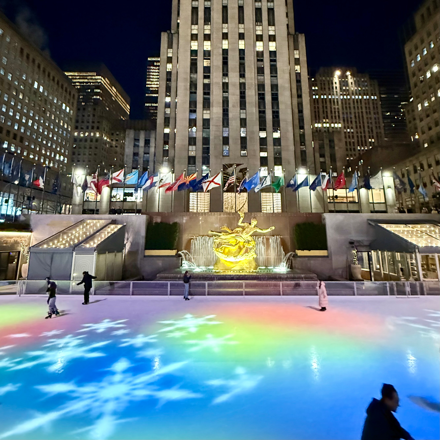 Auto-generated description: People are ice skating on a rink at Rockefeller Center, with colorful lights reflecting on the ice and a lit-up building in the background.