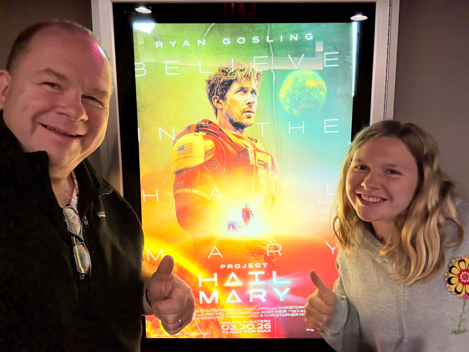 Man and teen girl giving thumbs up beside a lit Project Hail Mary movie poster showing Ryan Gosling in a spacesuit.