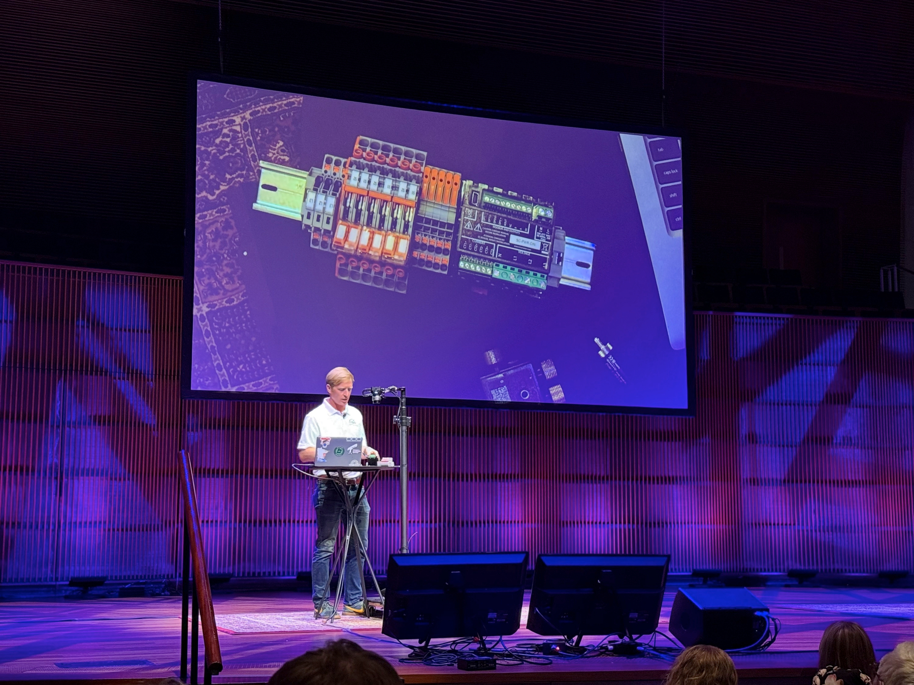 A presenter at a laptop on stage, with an overhead camera projecting a DIN rail of wired terminal blocks onto a large screen.
