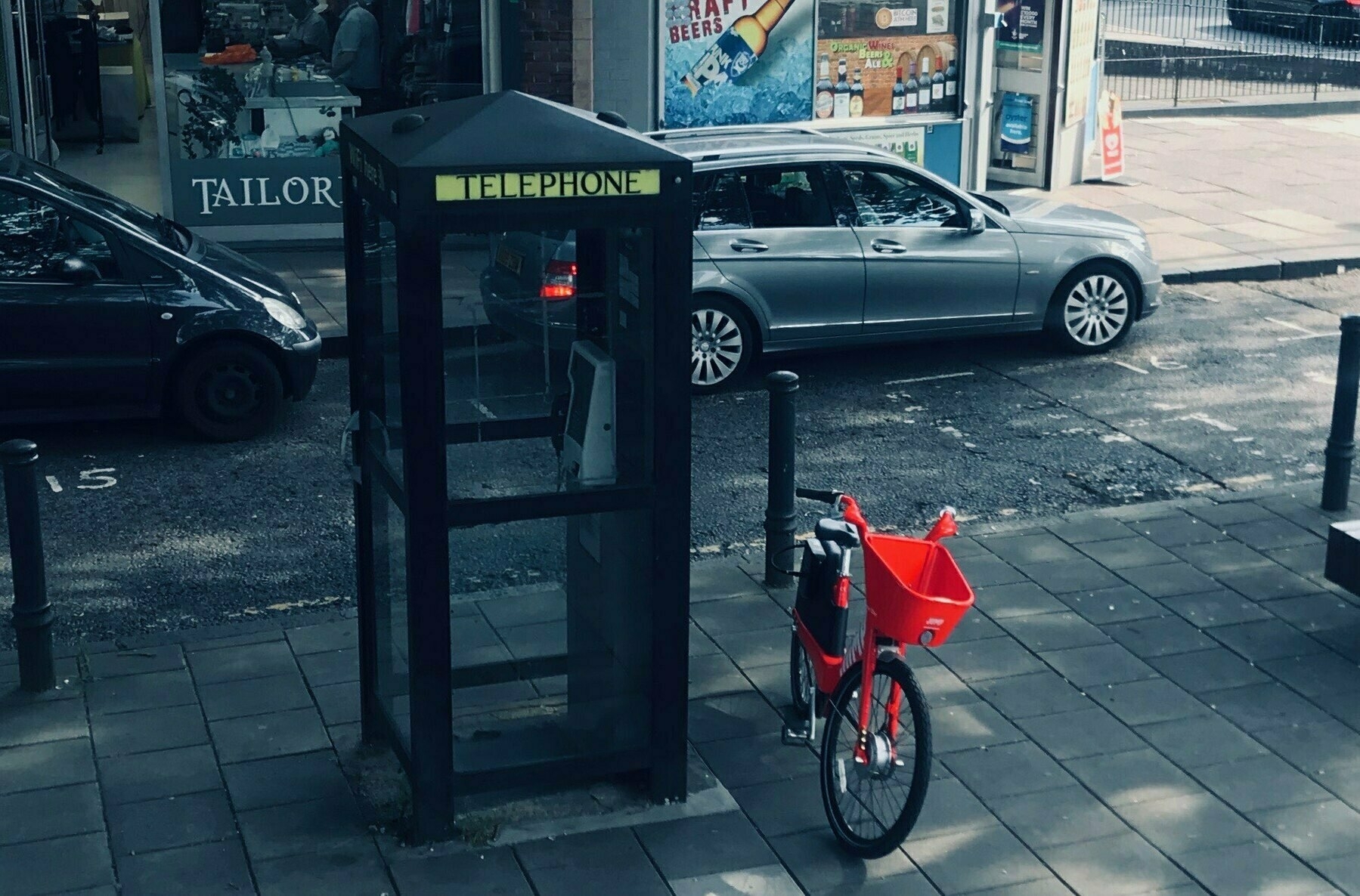 A bike with a bright red plastic front basket parked on a kickstand next a black telephone box on a London street