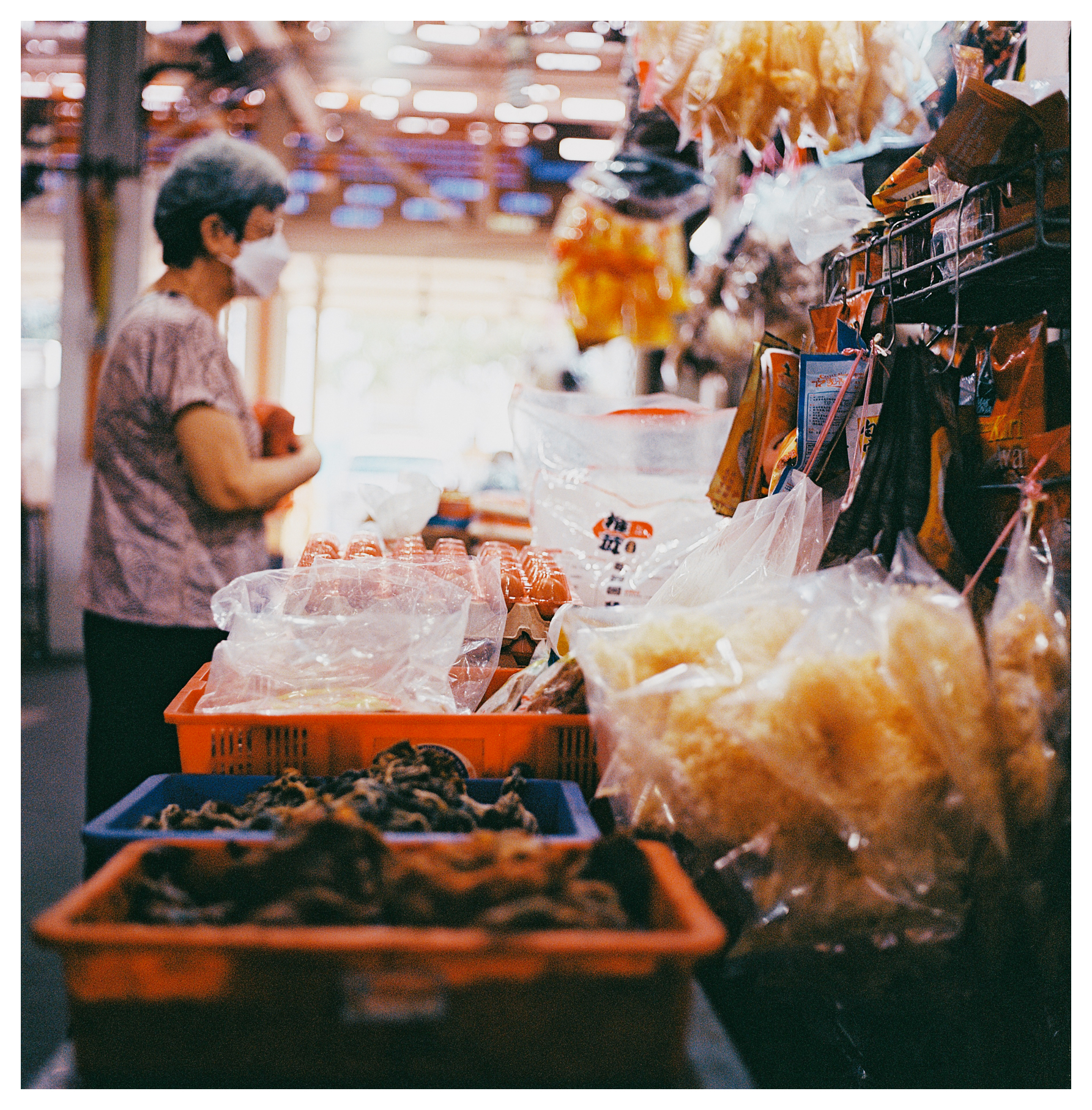 A scan of a medium format square color photo of an older Chinese lady wearing a face mask standing at a stall in Singapore looking at dried goods to purchase