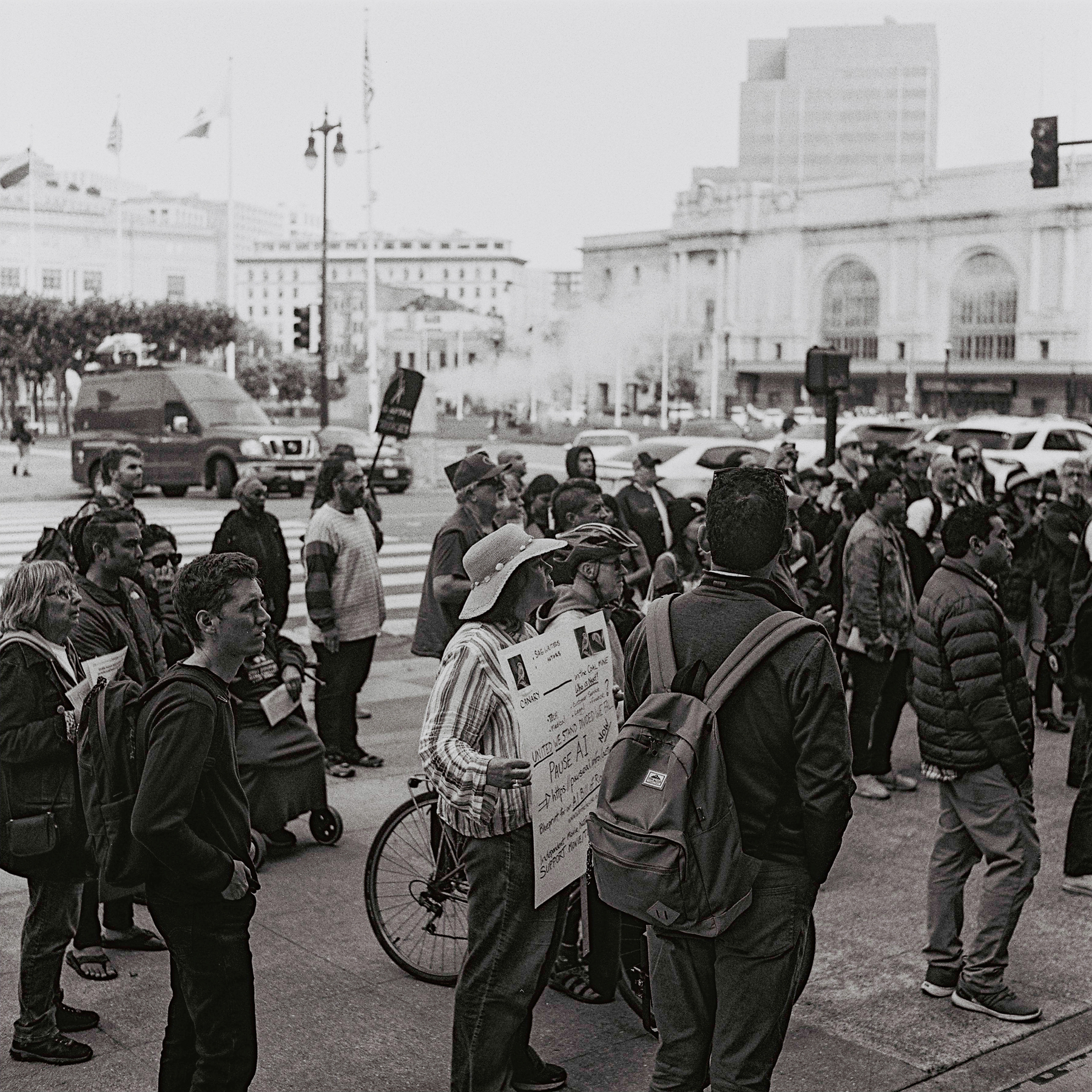 a scan of a black and white photo showing a gathering of people outside SF City Hall holding signs supporting the strike. In the background, half the photo has a scene of the field and Bill Graham auditorium building and a steam geyser with steam coming out of the road