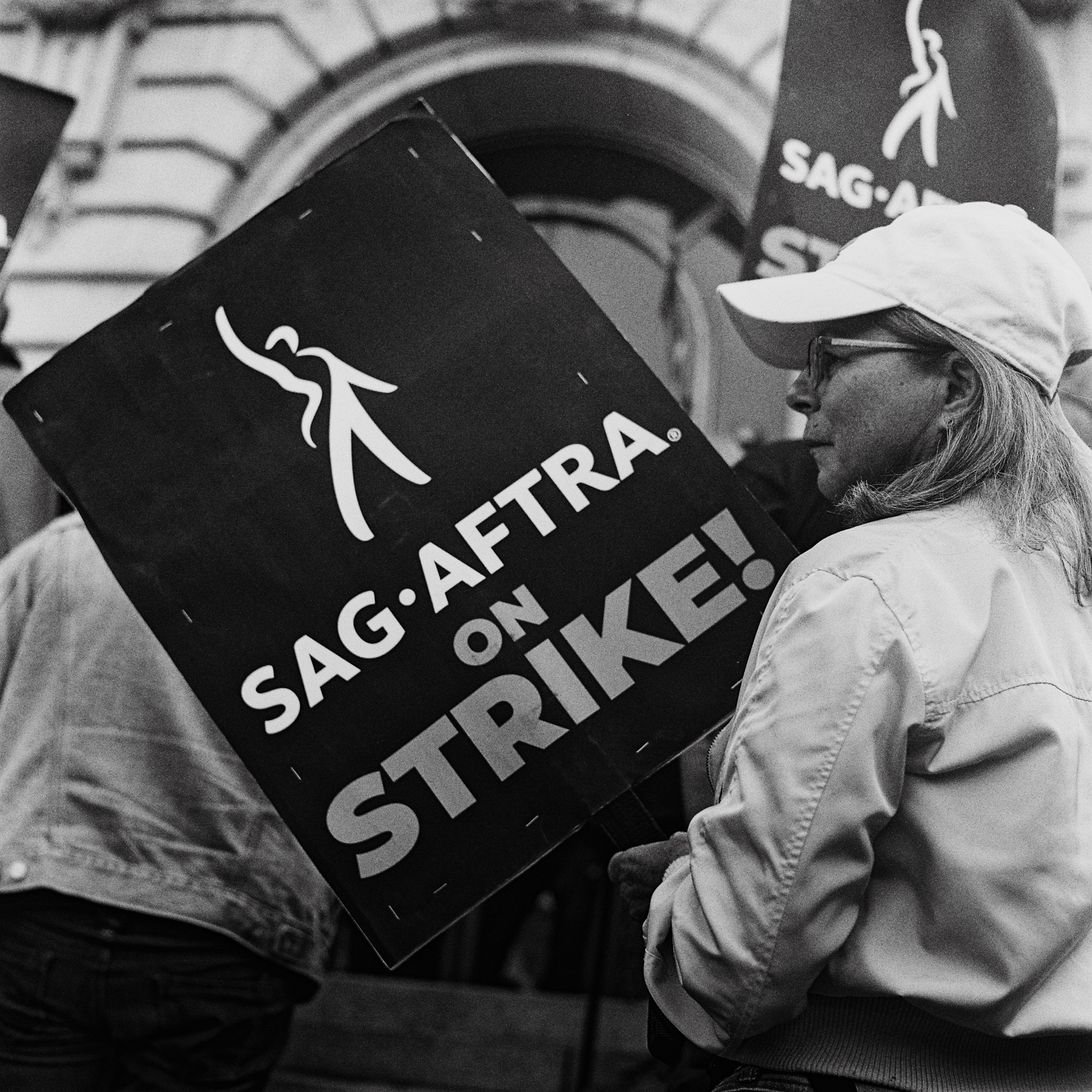 a scan of a black and white photo showing a woman holding a SAG-AFTRA on Strike placard, she has her side view of her face turned to the camera and she looks serious