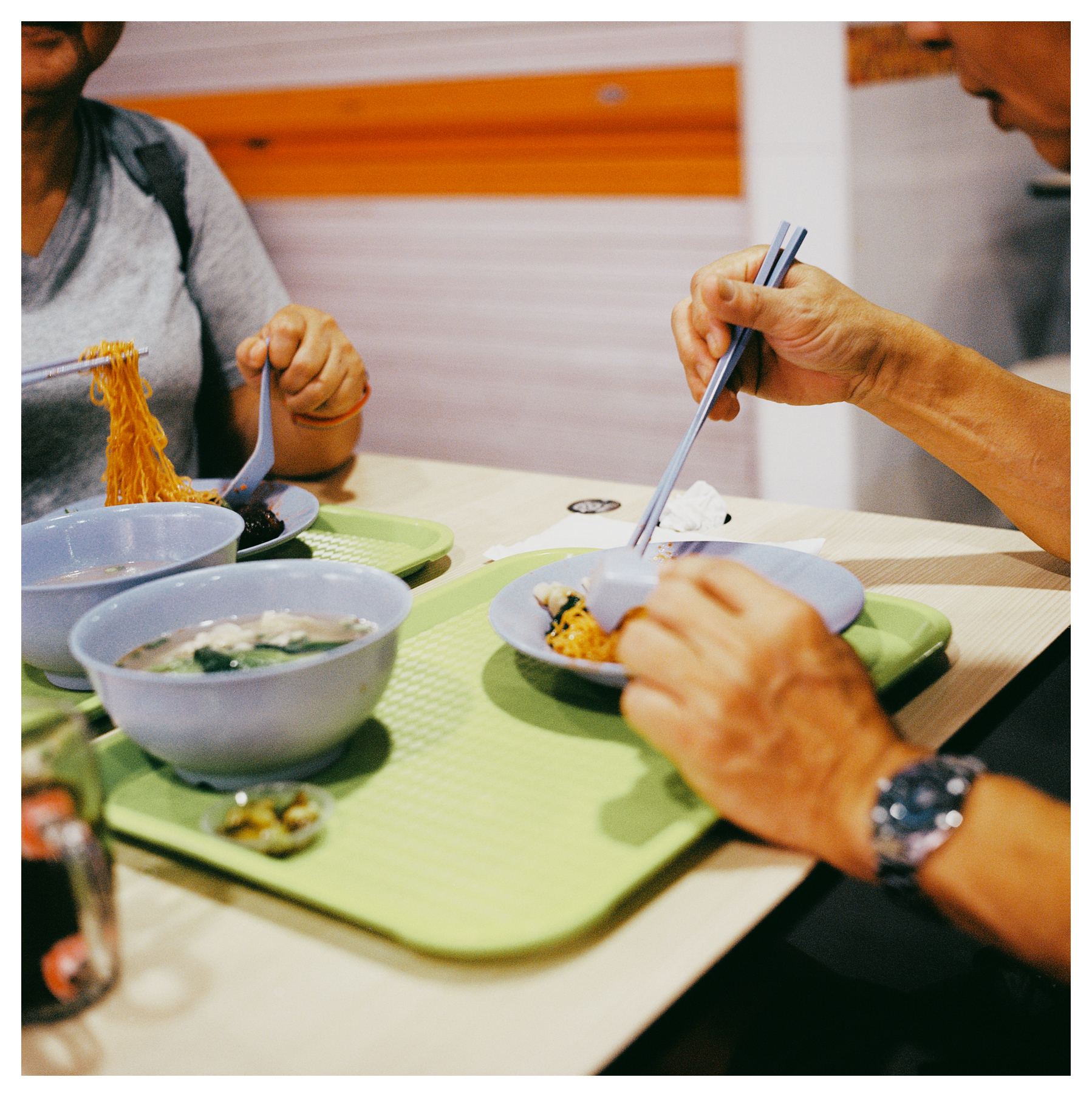 A scan of a medium format square color photo of two older Chinese people eating noodles with chopsticks