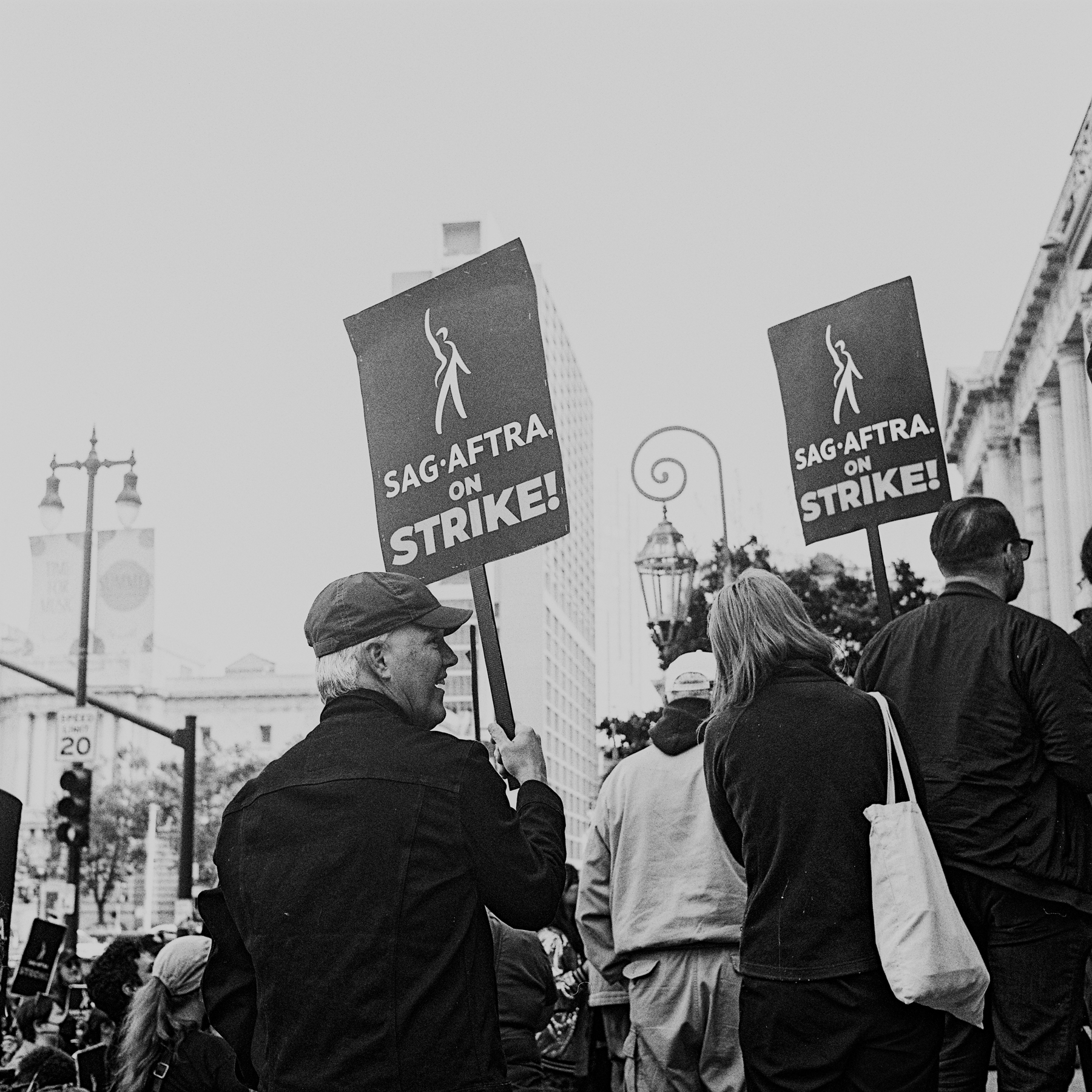 a scan of a black and white photo showing a man holding a sign that says SAG-AFTRA on strike, with his side profile turned towards the camera. he is smiling