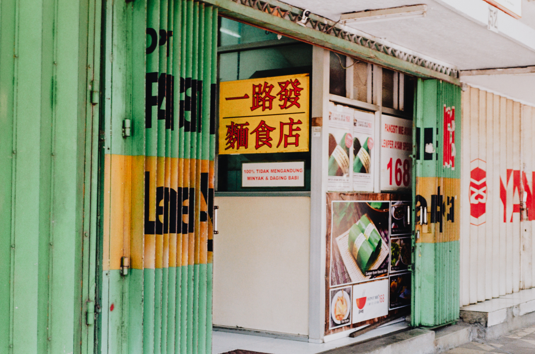 a scan of a color photo showing a Chinese Indonesian noodle shop in old Surabaya with Chinese and Indonesian words. The door is green with yellow features