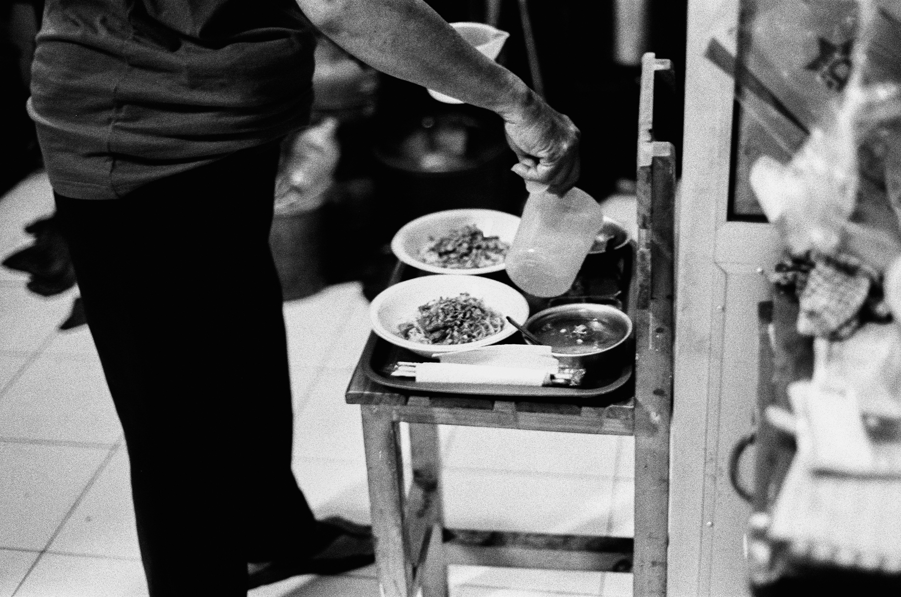 a scan of a black and white photo of a person's feet, hovering near where there are noodles and soup being prepared on a tray