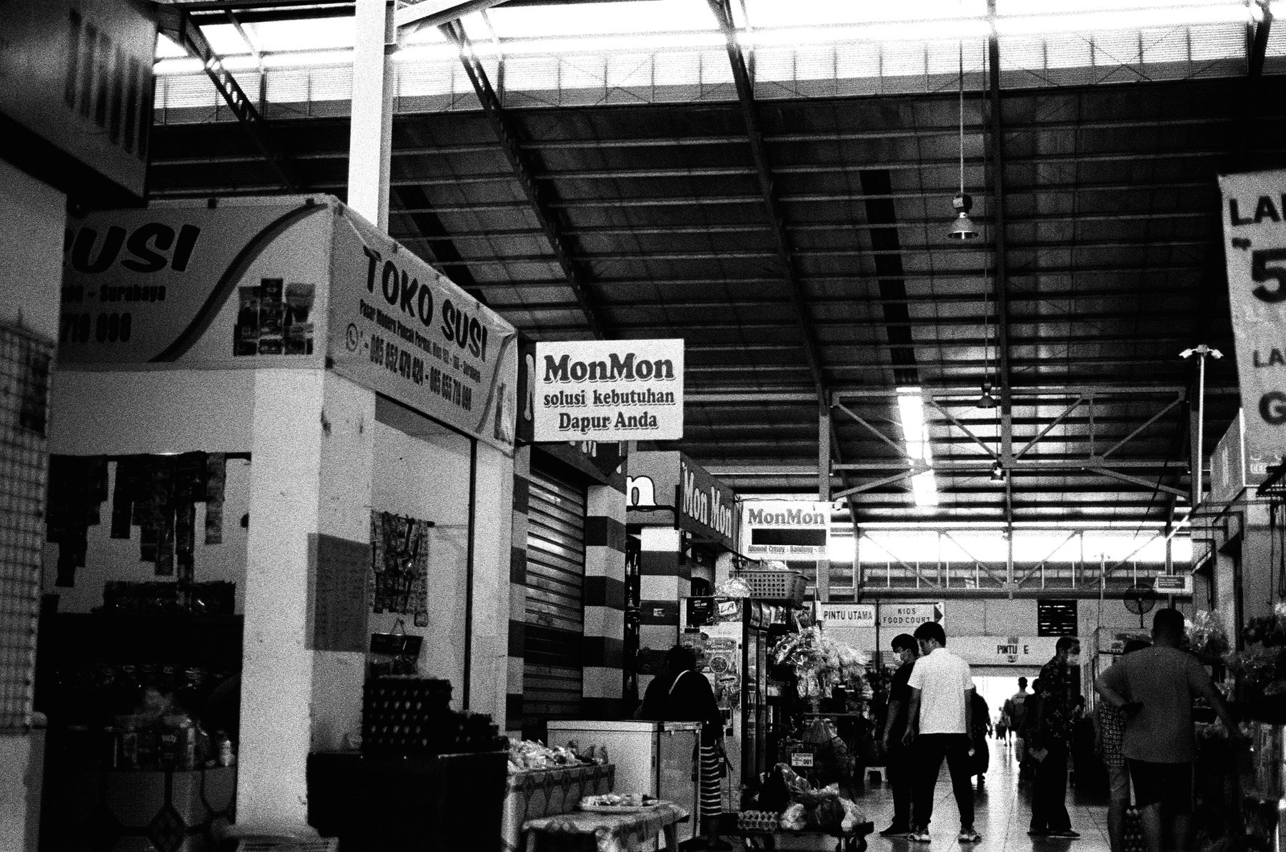 a scan of a black and white photo of the interior of a wet market in indonesia, showing many shops with indonesian signs