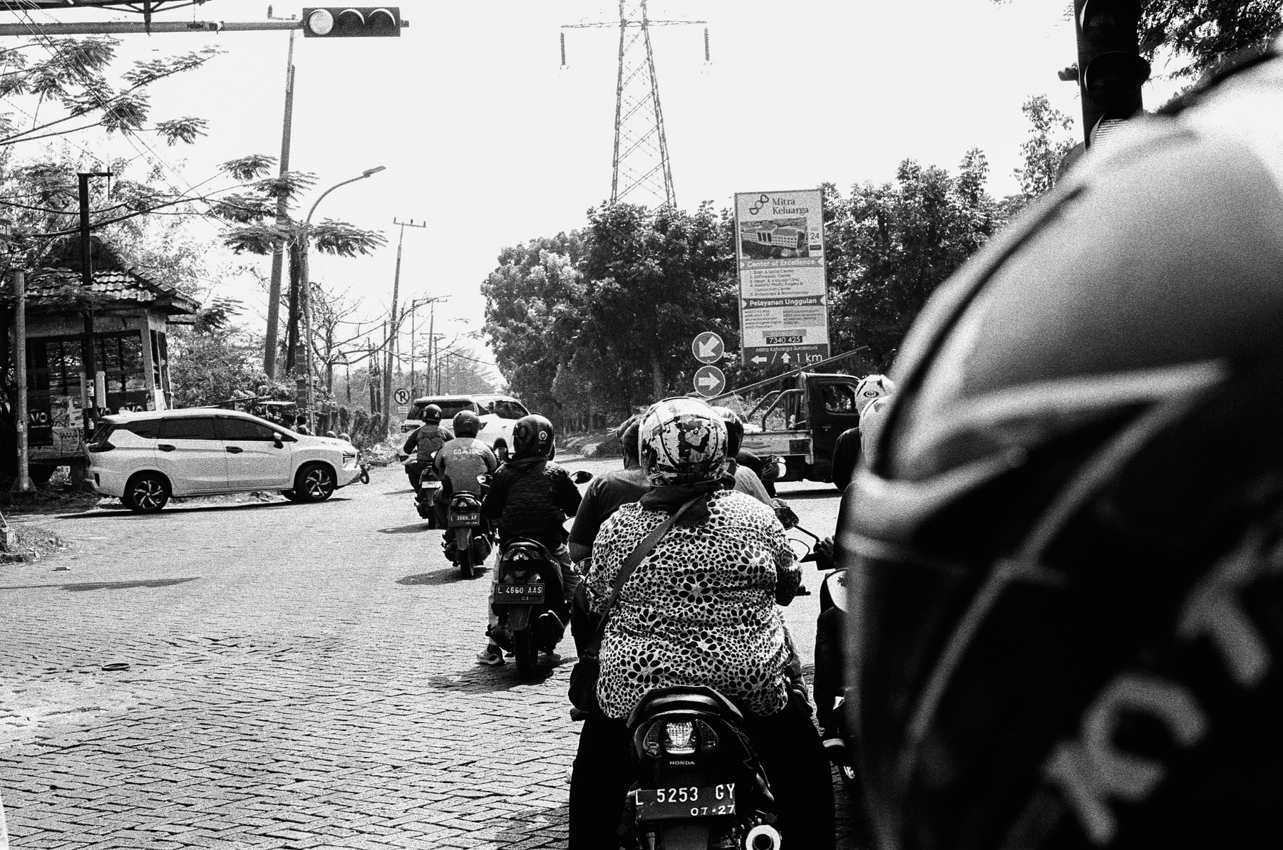 a scan of a black and white photo of traffic in surabaya from the back of a motorbike, pillion. in front, several motorbikes and their passengers stopped at traffic