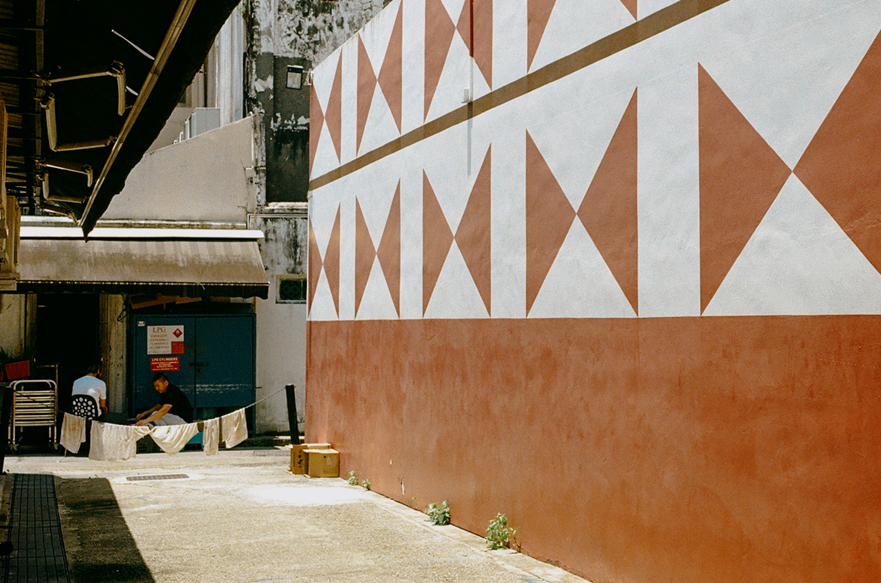 5. A scan of a color photo of two men in little India sitting at the back of a restaurant. An alley opens up into them, and the foreground wall is painted with geometric clay color and shapes, indicating it is part of a Hindu temple next door