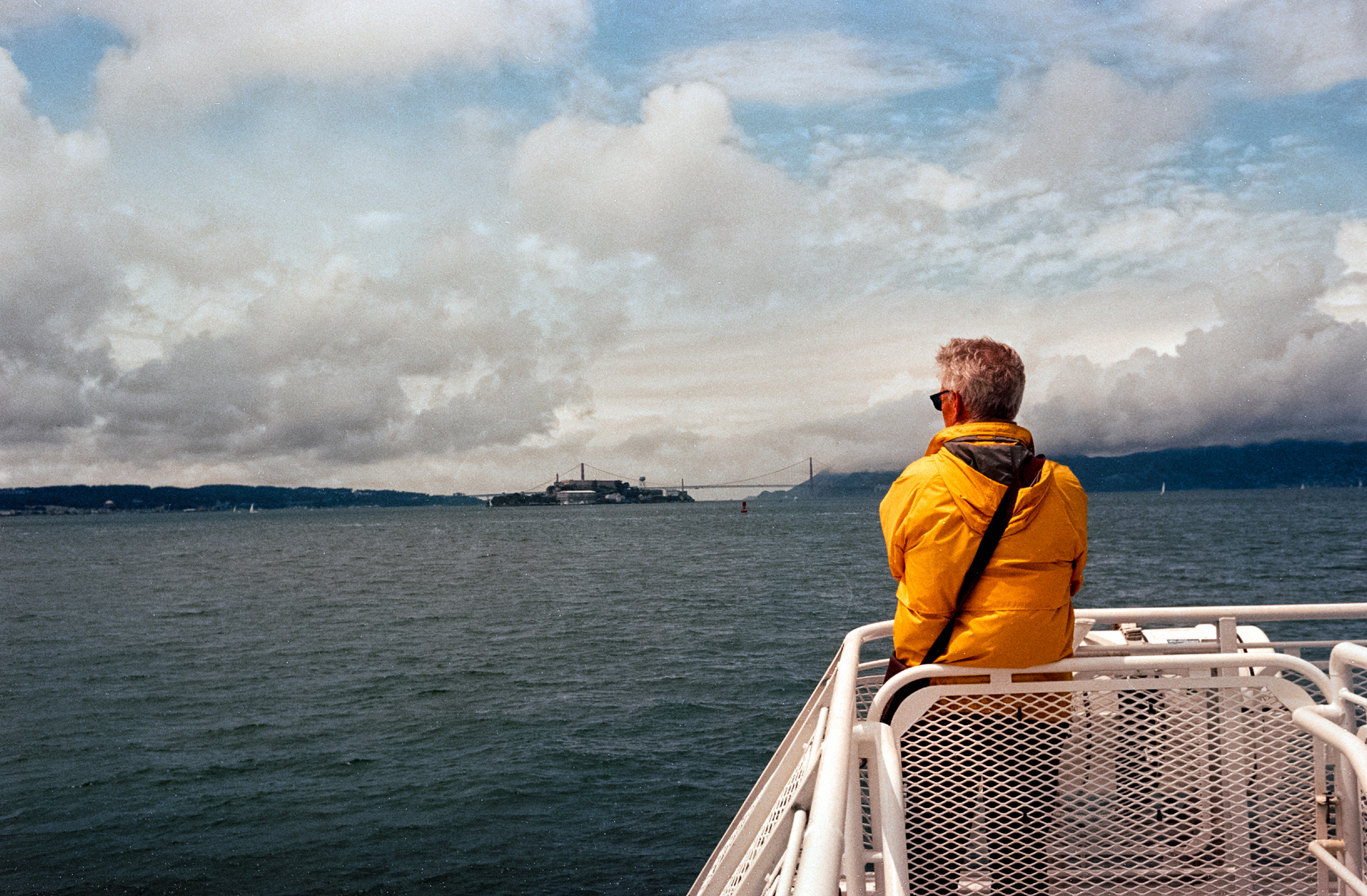 A scan of a color photograph of a man on a ferry, wearing a yellow jacket looking at Alcatraz in the distance