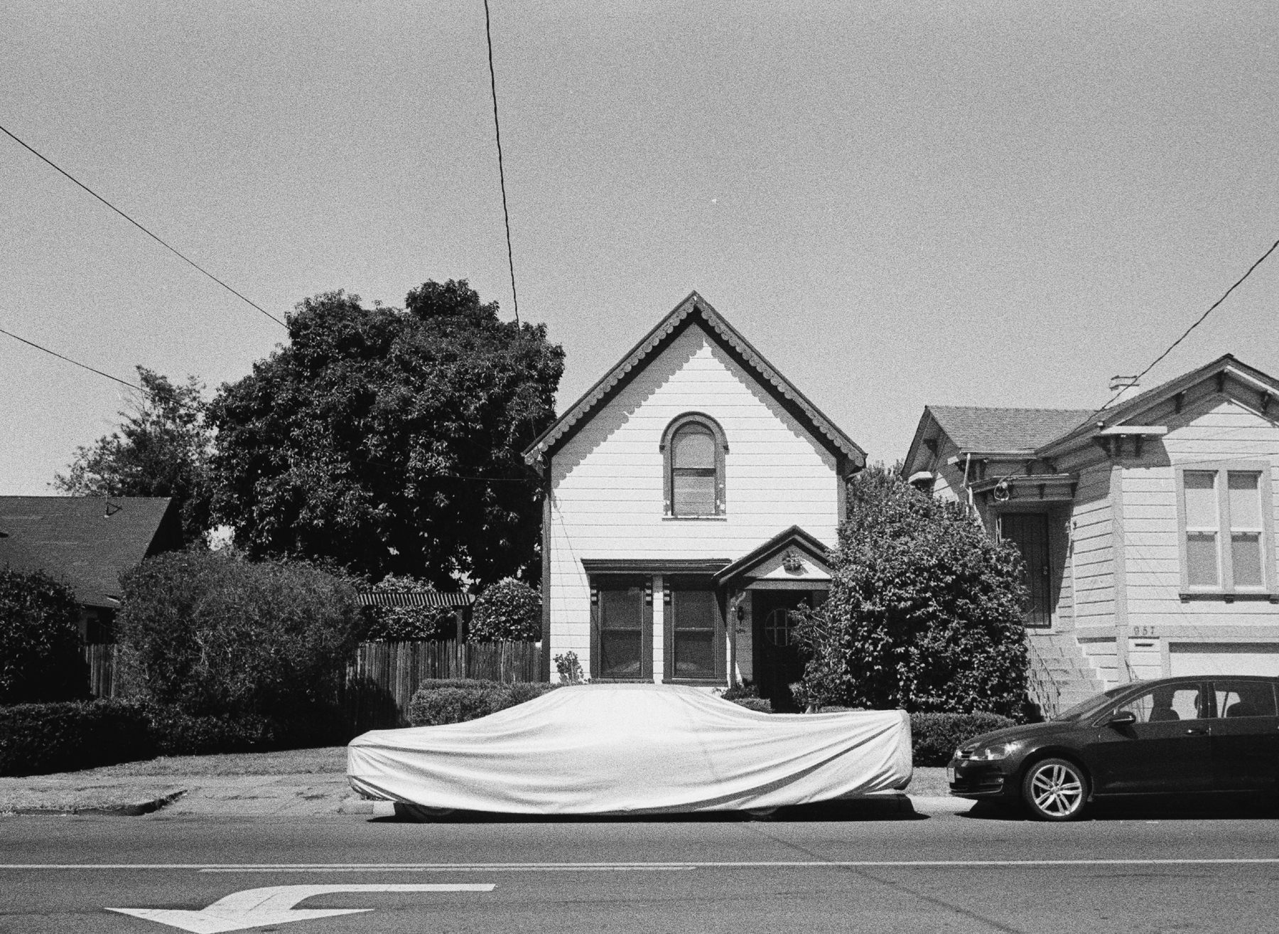 a scan of a black and white photo showing an old school home with a car covered with a sheet outside, the sheet has a clear outline of a retro style car.