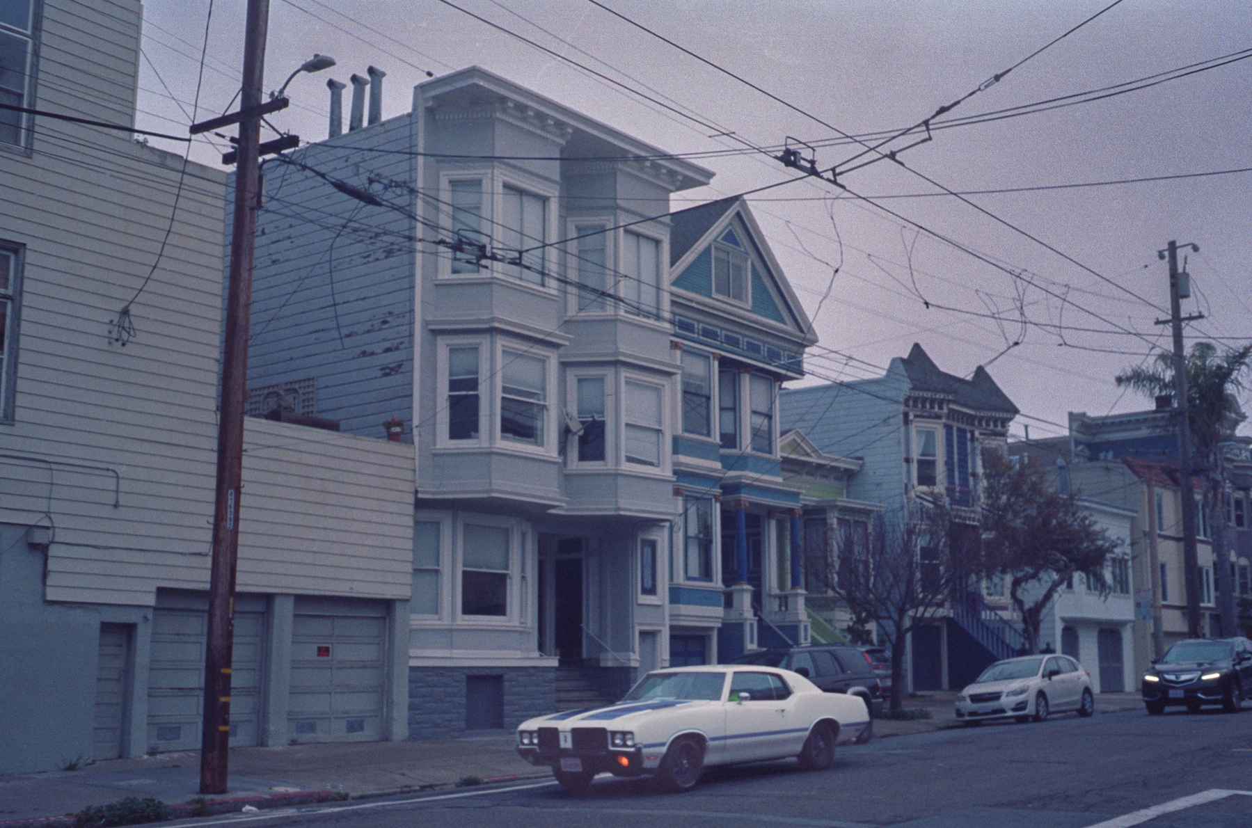 a scan of a color photograph of a neighborhood in Cole Valley San Francisco where the Victorian houses are mostly blue and there is a retro white car parked on the street