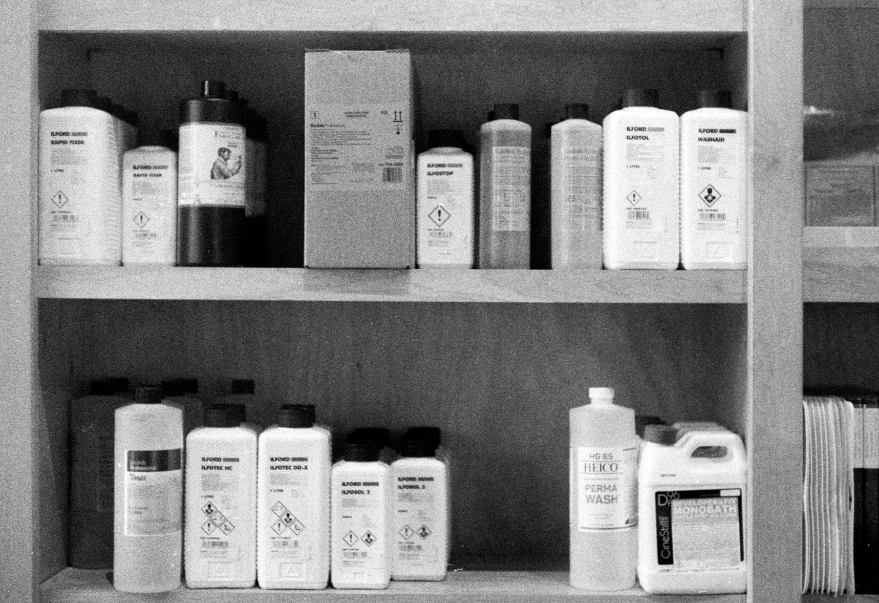 a scan of a black and white photo showing some film development chemicals on a shelf in a photography store, brands include Ilford and Kodak