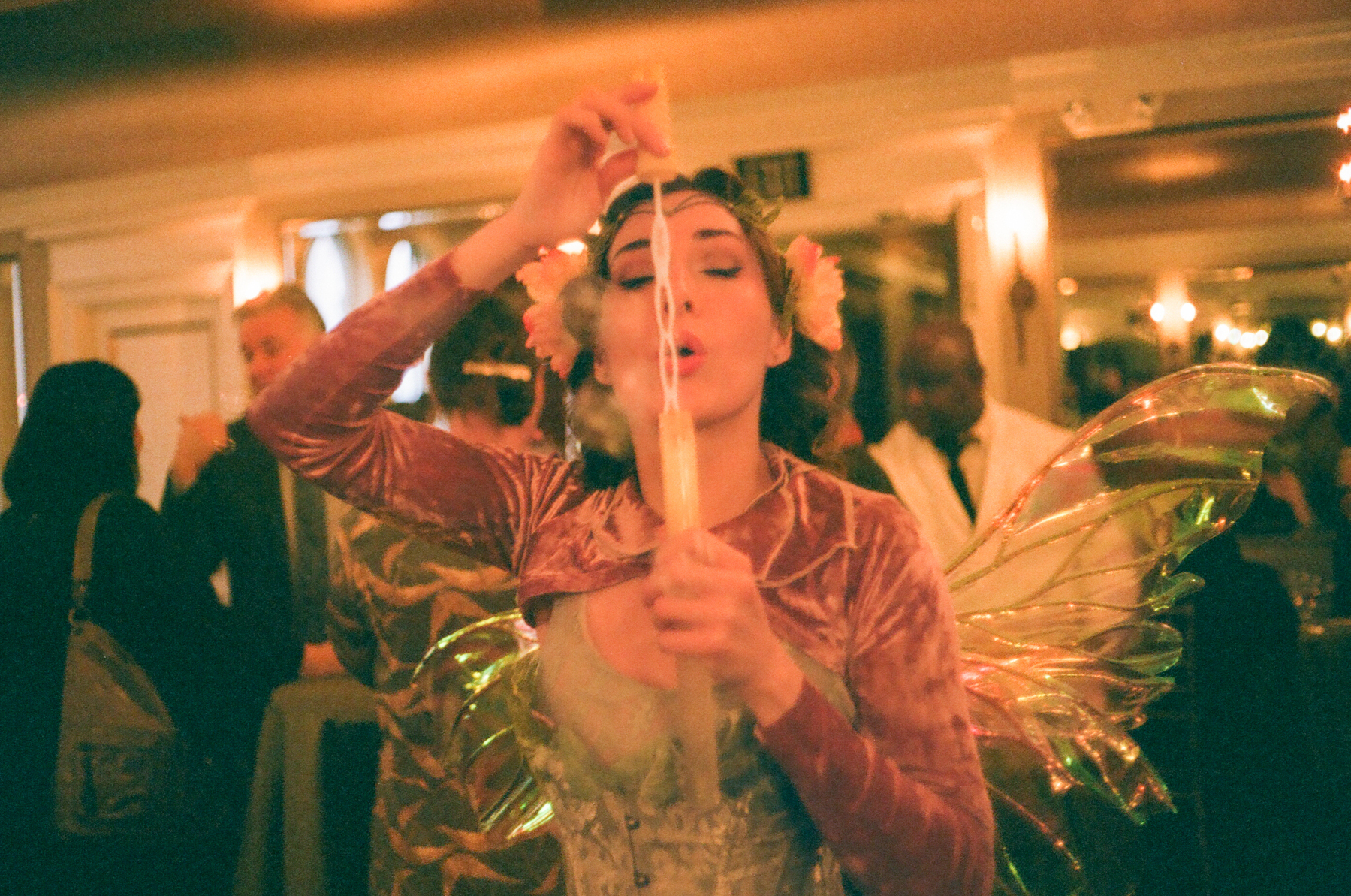 a scan of a color photograph showing a beautiful woman dressed as a fairy, blowing bubbles at the camera, in a ballroom-like setting filled with people wearing formal attire