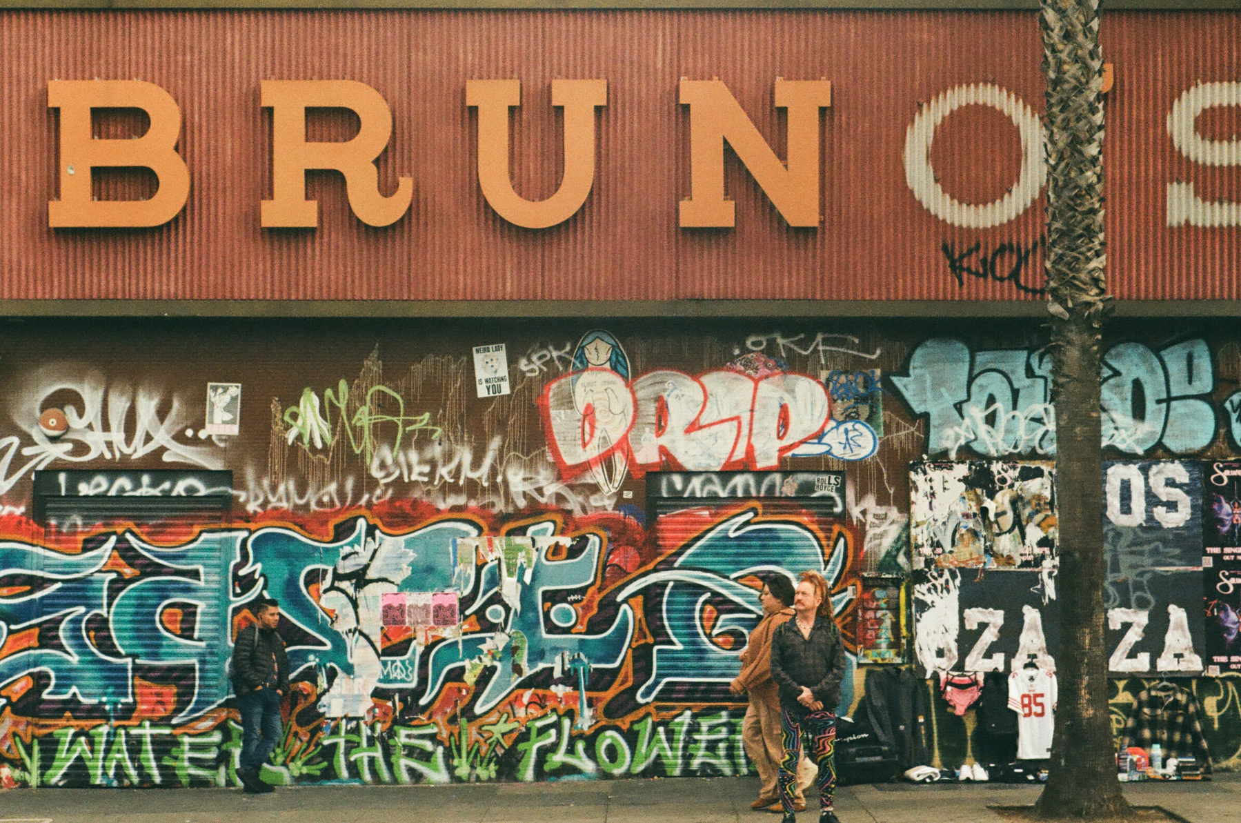 A color photograph of a street scene in the Mission district, San Francisco, with a large sign that says Bruno's and a few people standing in front of a heavily graffitied wall