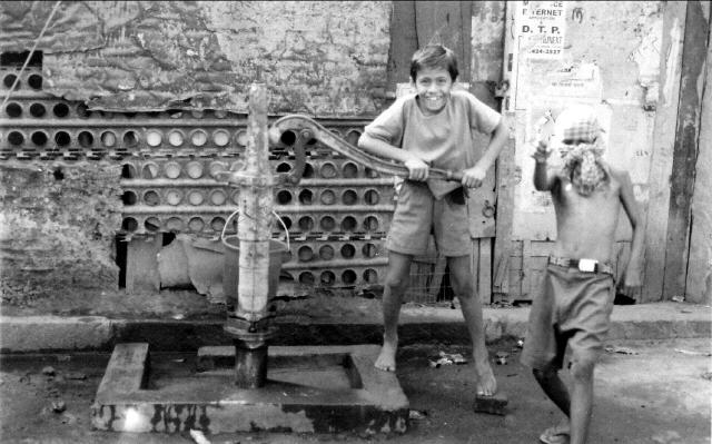 A black and white photo of two boys horsing around at a water pump in Kolkata, India